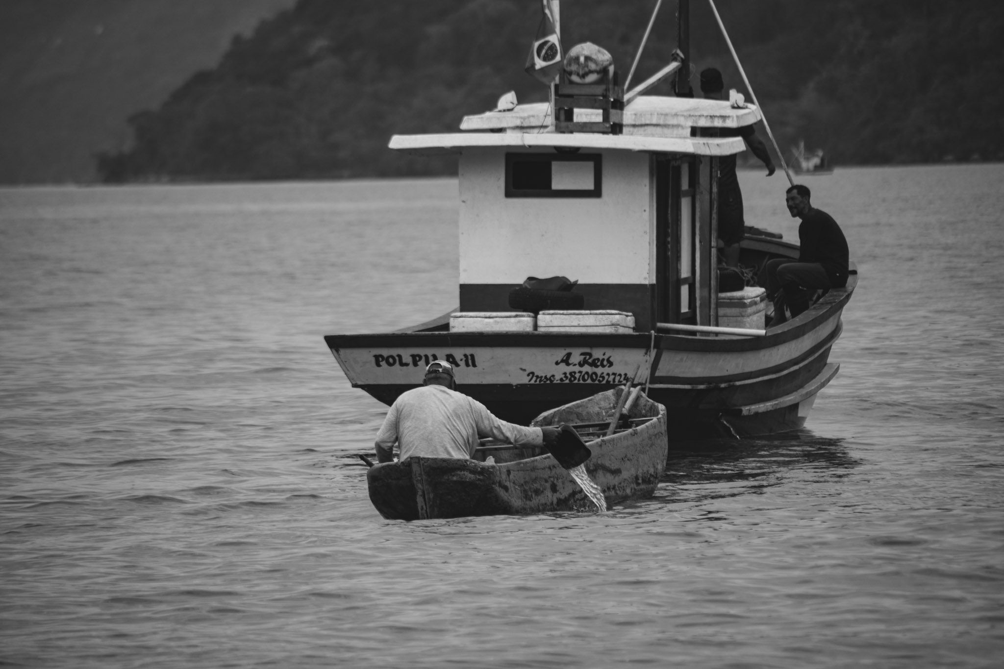 Hombre remando en un bote chico hacia un barco pesquero, en blanco y negro