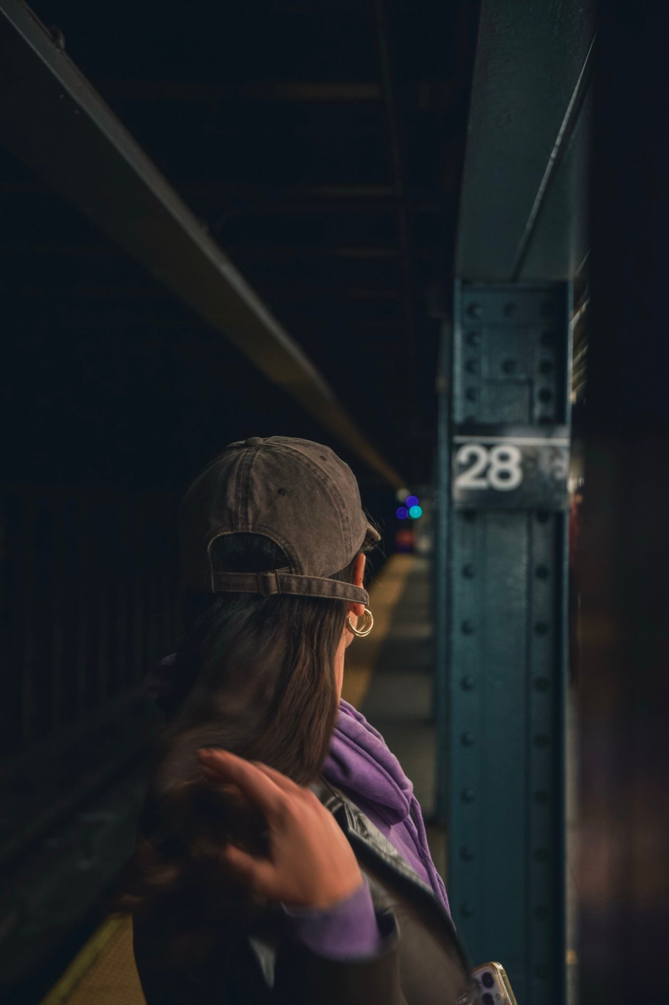 Una chica de espaldas con gorra y buzo violeta en el andén del subte, junto a una columna con el número 28