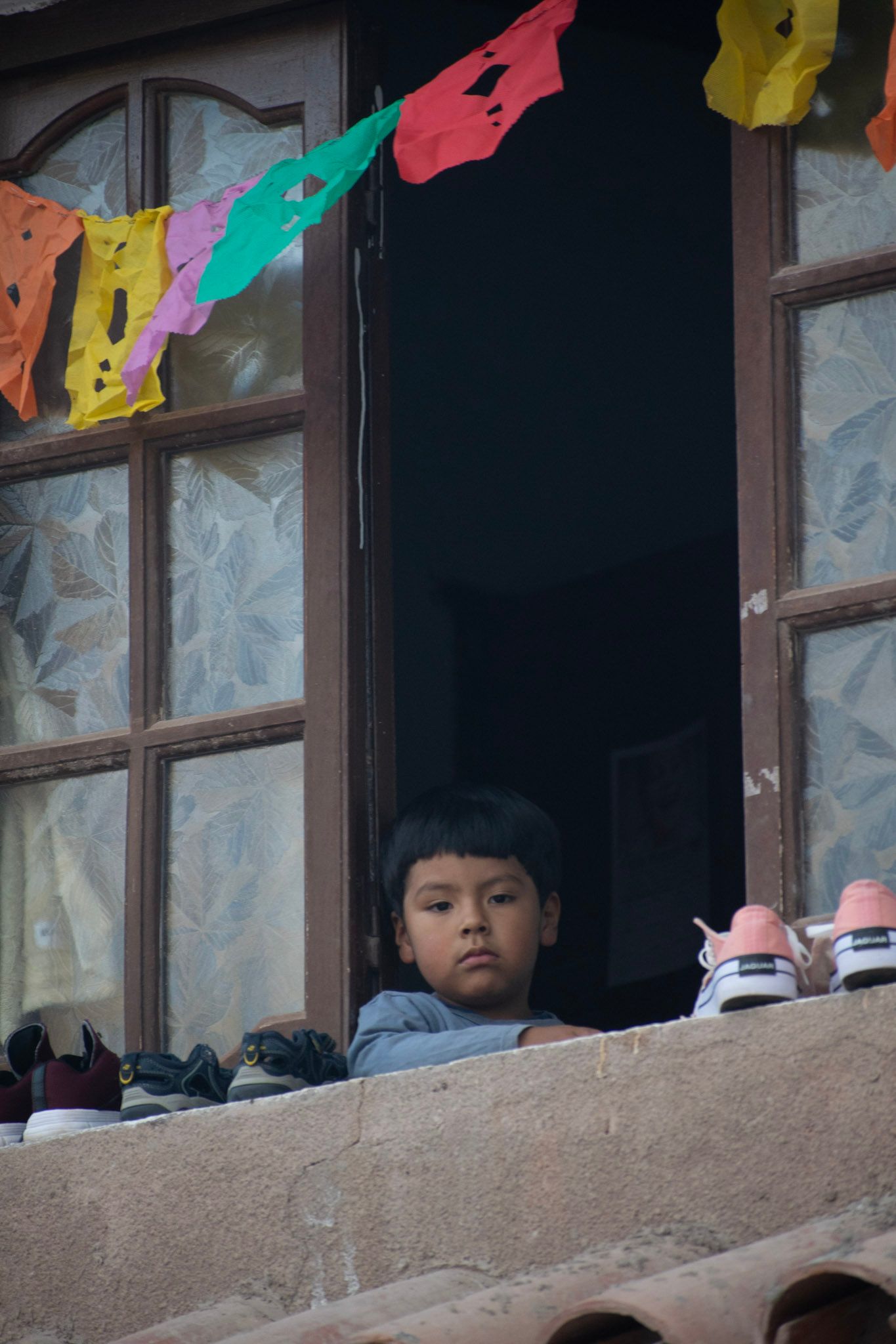 Nene asomado a una ventana con zapatillas puestas a secar en el alféizar y banderines de papel picado de colores