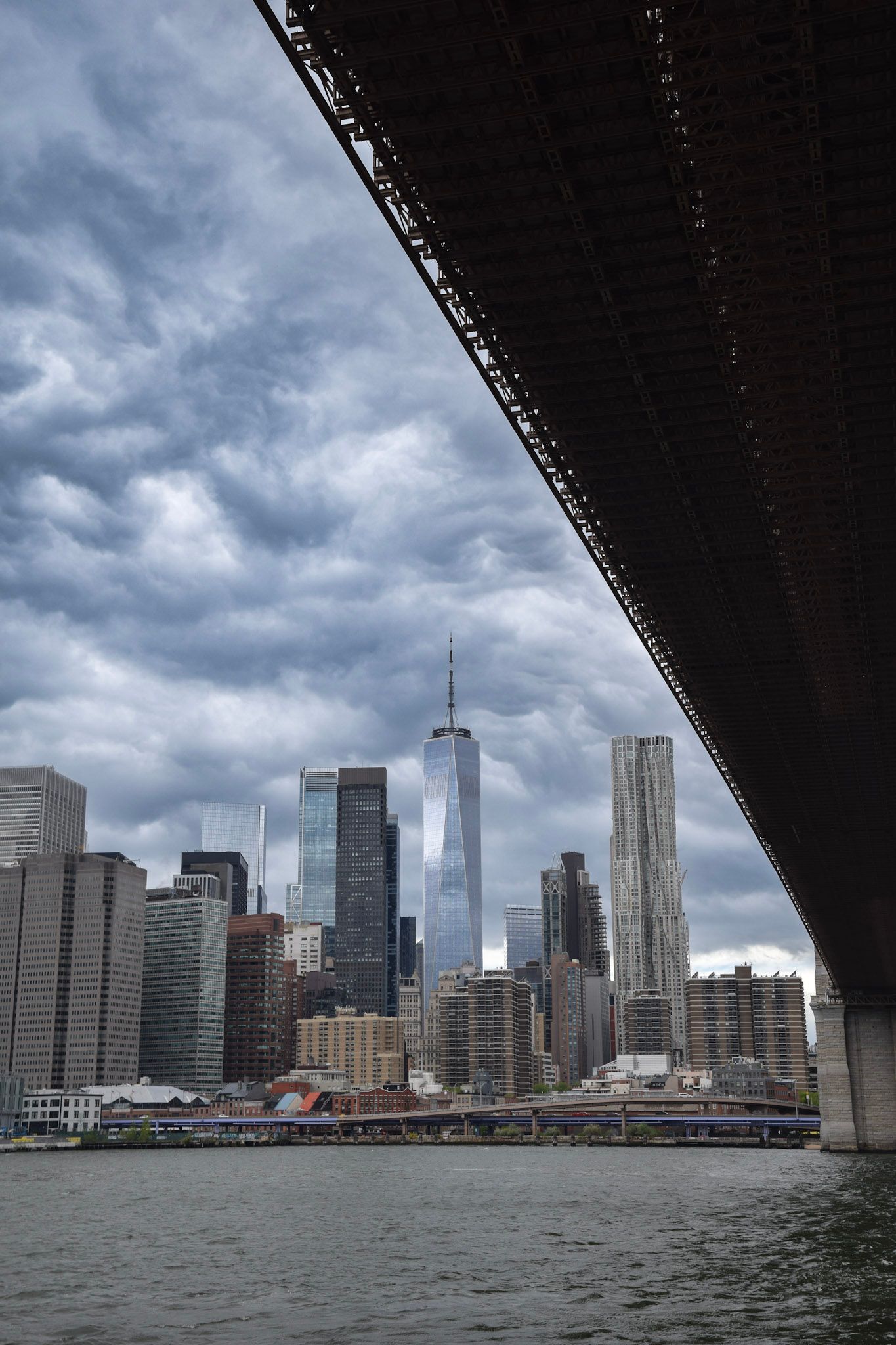 Los rascacielos del Lower Manhattan y el One World Trade Center bajo un cielo de nubes grises, vistos desde el río debajo del puente de Brooklyn