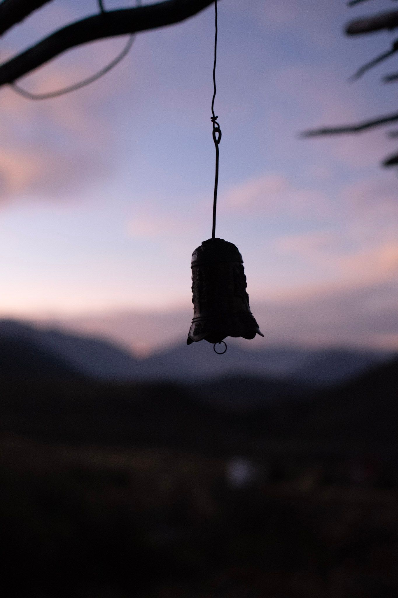 Campana de metal colgando de un alambre en silueta, contra un cielo rosado y montañas desenfocadas al atardecer