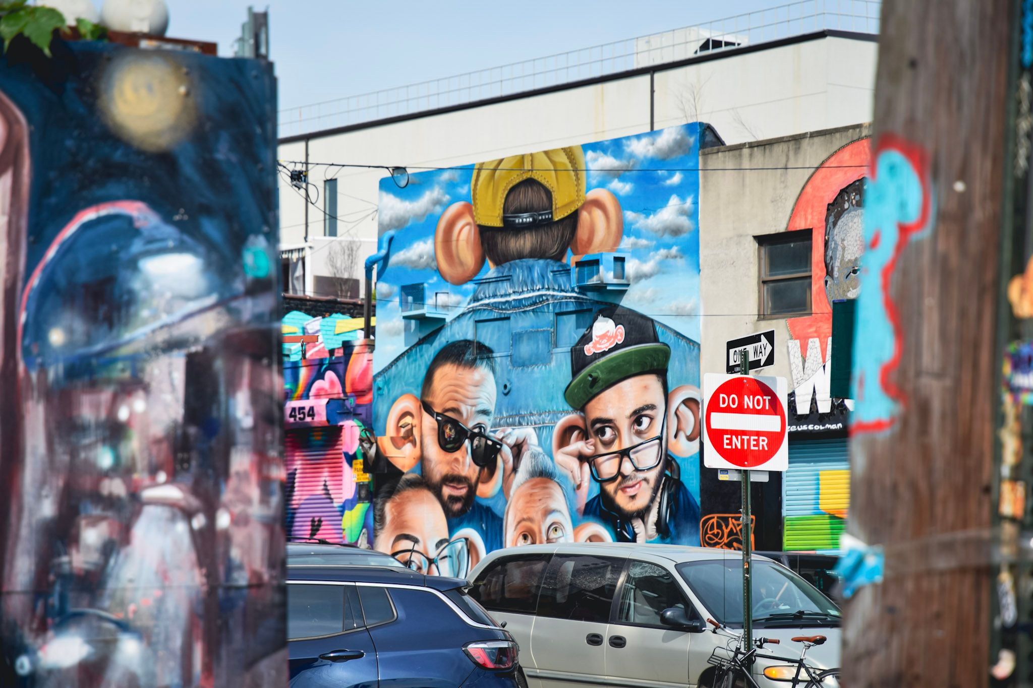 Mural de caras caricaturescas con gorras y anteojos sobre un edificio, con autos estacionados y un cartel de contramano