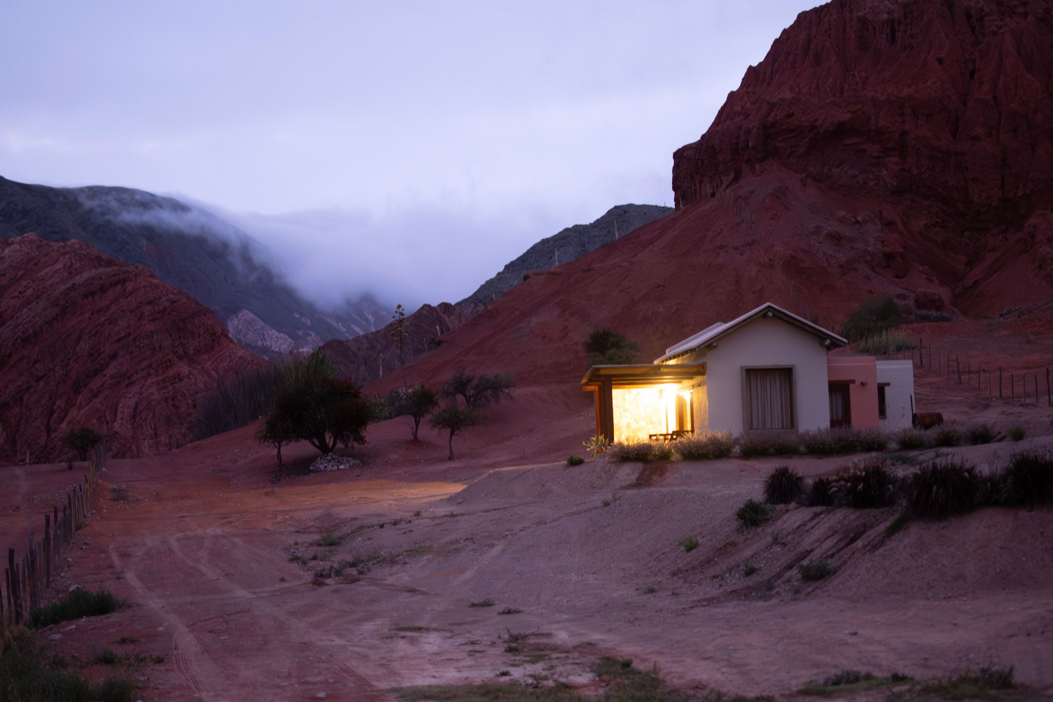 Casa sola con la luz encendida al pie de cerros rojos, al anochecer, con nubes bajas entre las montañas