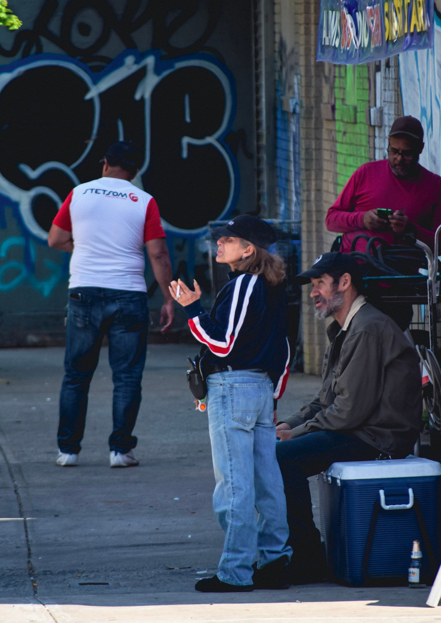 Una mujer con gorra gesticula mientras habla con un hombre de barba sentado junto a una conservadora, frente a una pared con grafitis