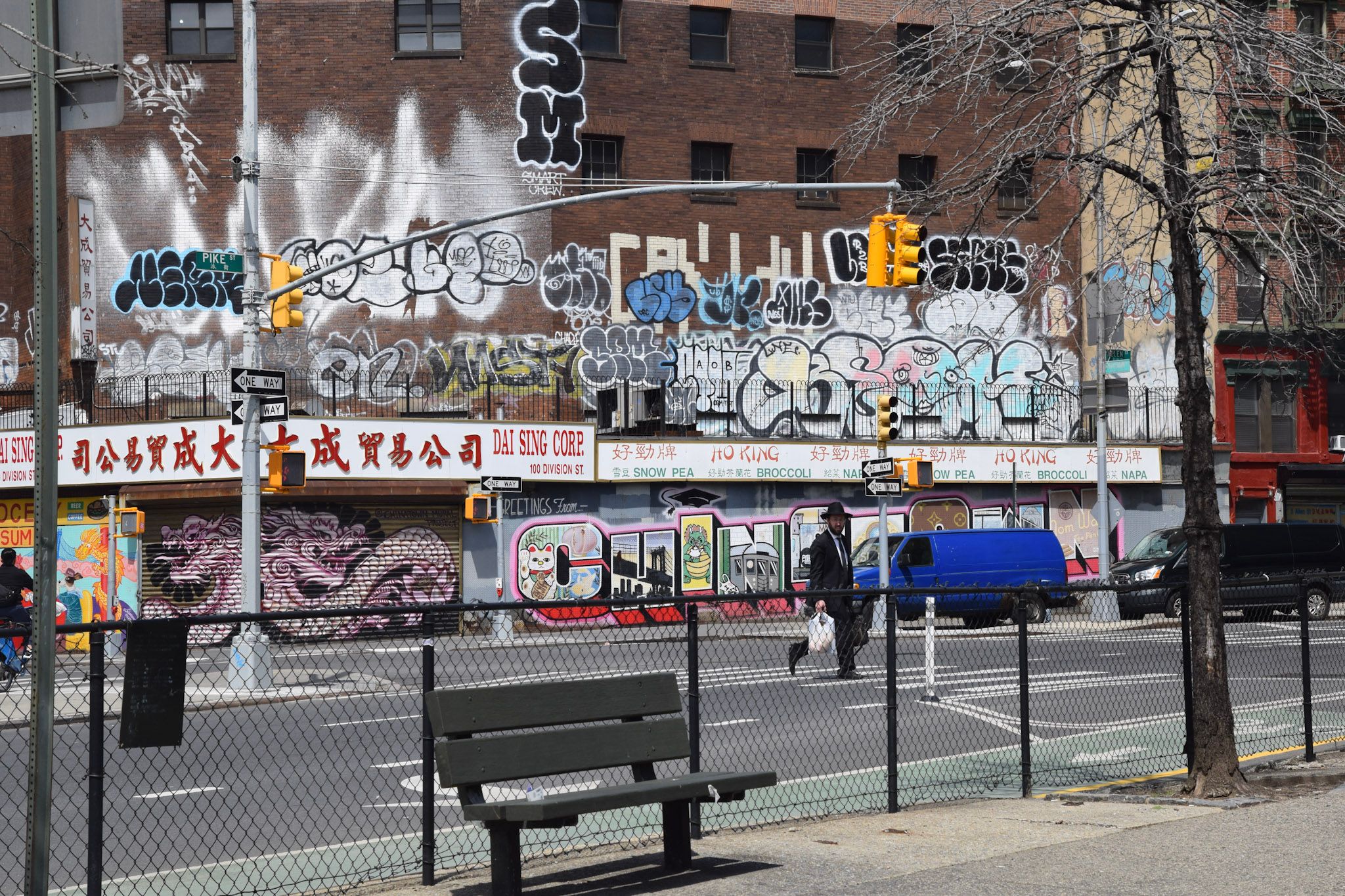 Pared cubierta de grafitis sobre comercios con carteles en chino, con un banco y un alambrado en primer plano