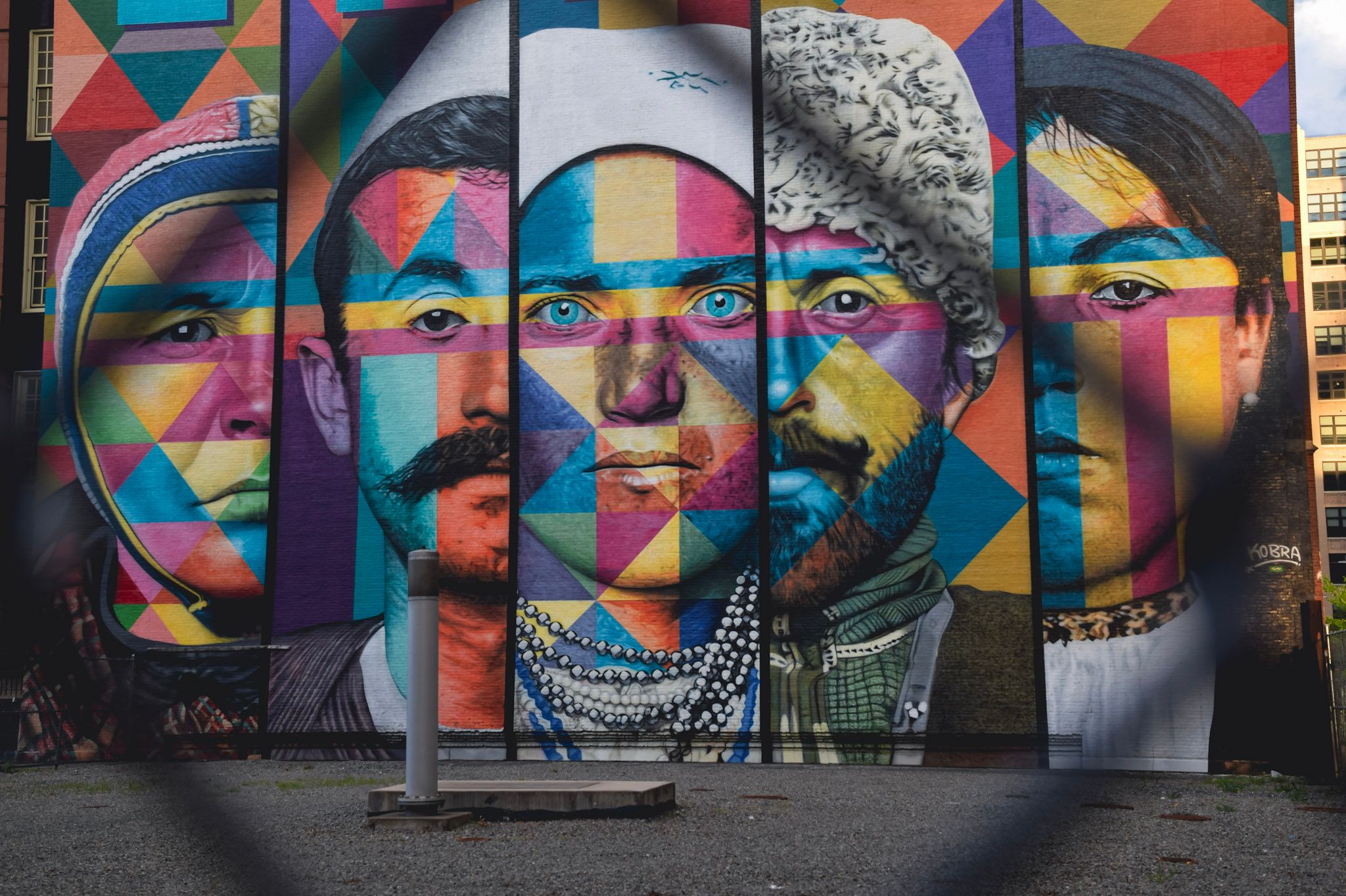Mural de cinco retratos con trajes tradicionales pintados con triángulos de colores, visto a través de un alambrado