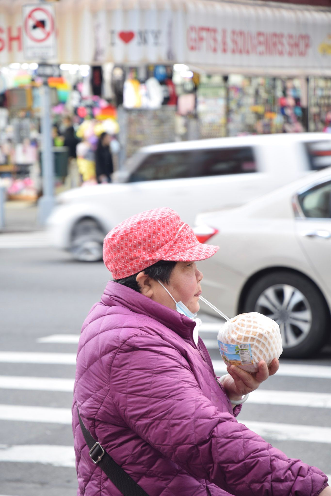 Una mujer con gorra roja y campera violeta toma de un coco con un sorbete, cruzando la calle frente a una tienda de souvenirs