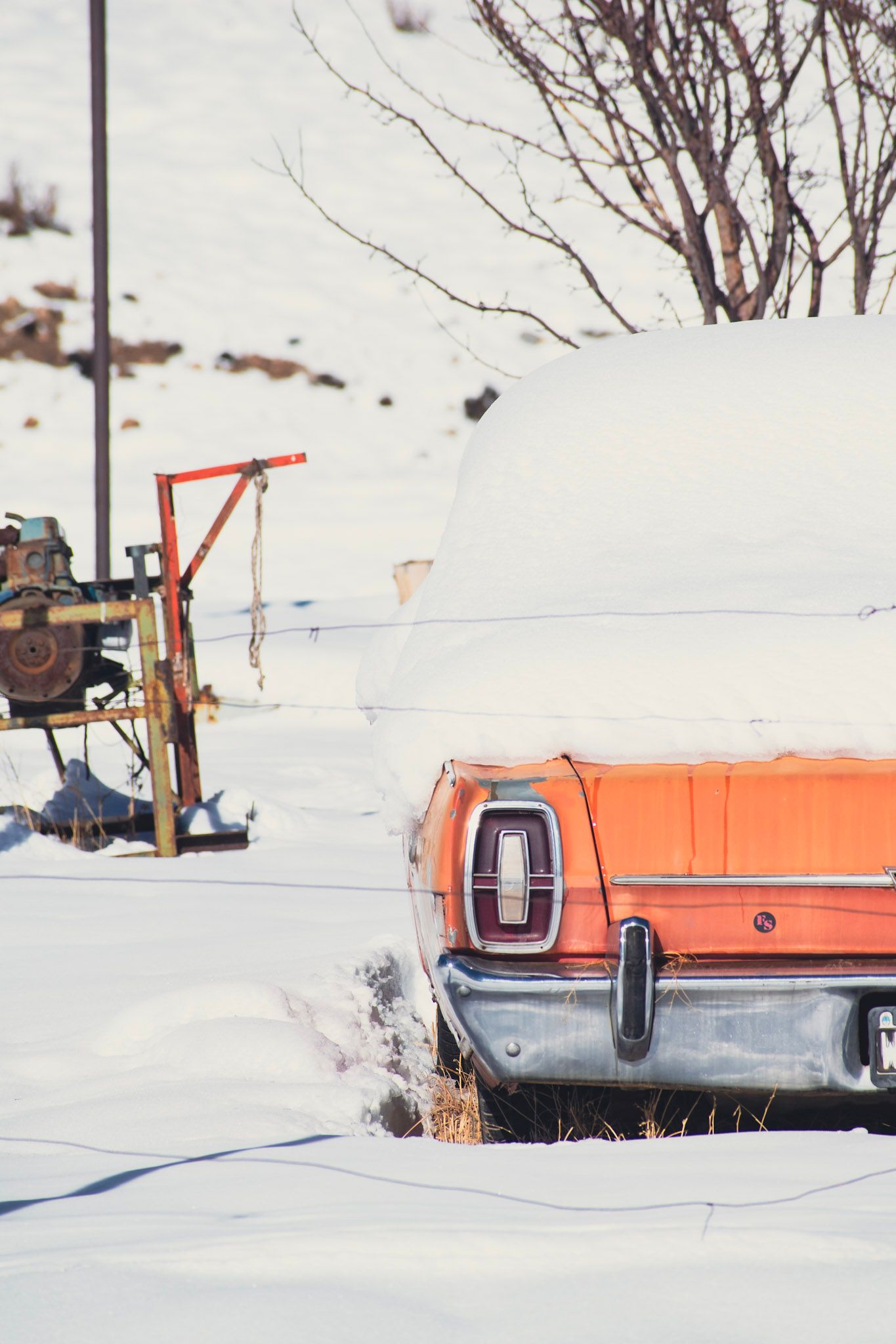 Parte trasera de un auto viejo naranja tapado de nieve, junto a una máquina oxidada