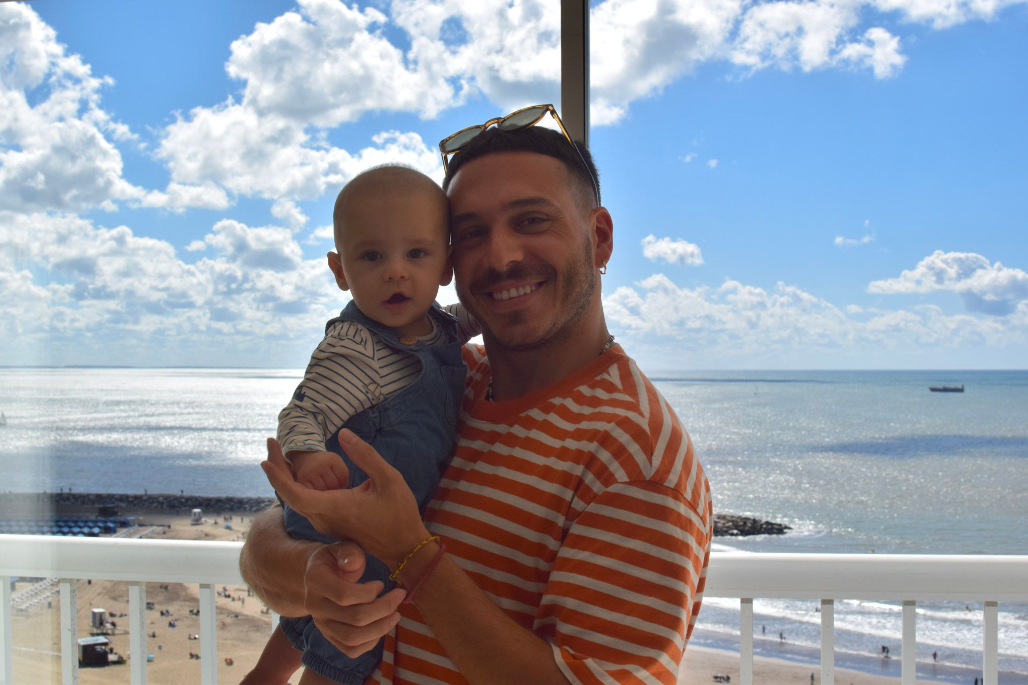 Mauruch sonriendo con su hijo en brazos en un balcón, con el mar y un cielo con nubes de fondo