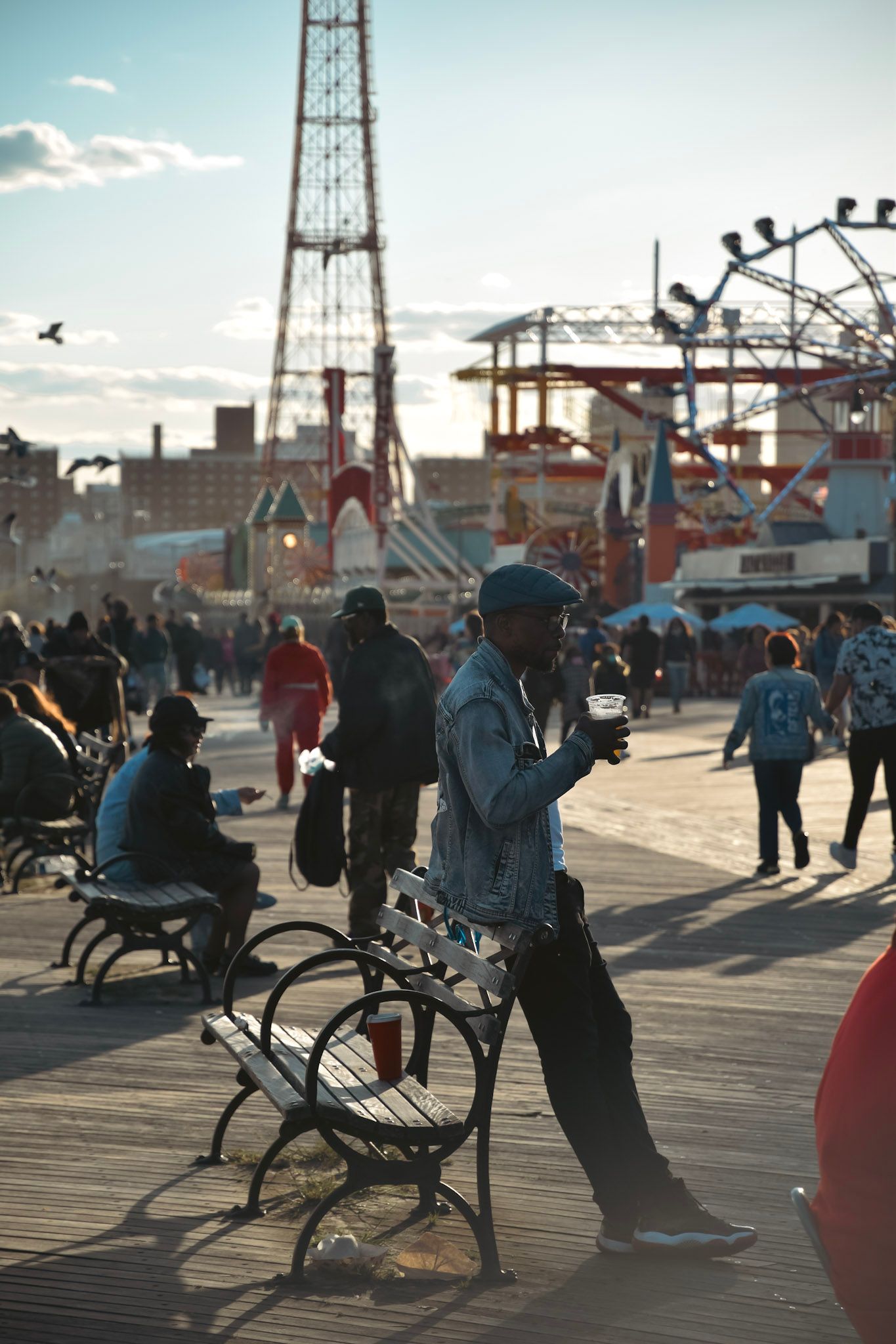 Un hombre con gorra y campera de jean apoyado en un banco de la rambla de Coney Island, con un vaso en la mano y los juegos del parque de fondo
