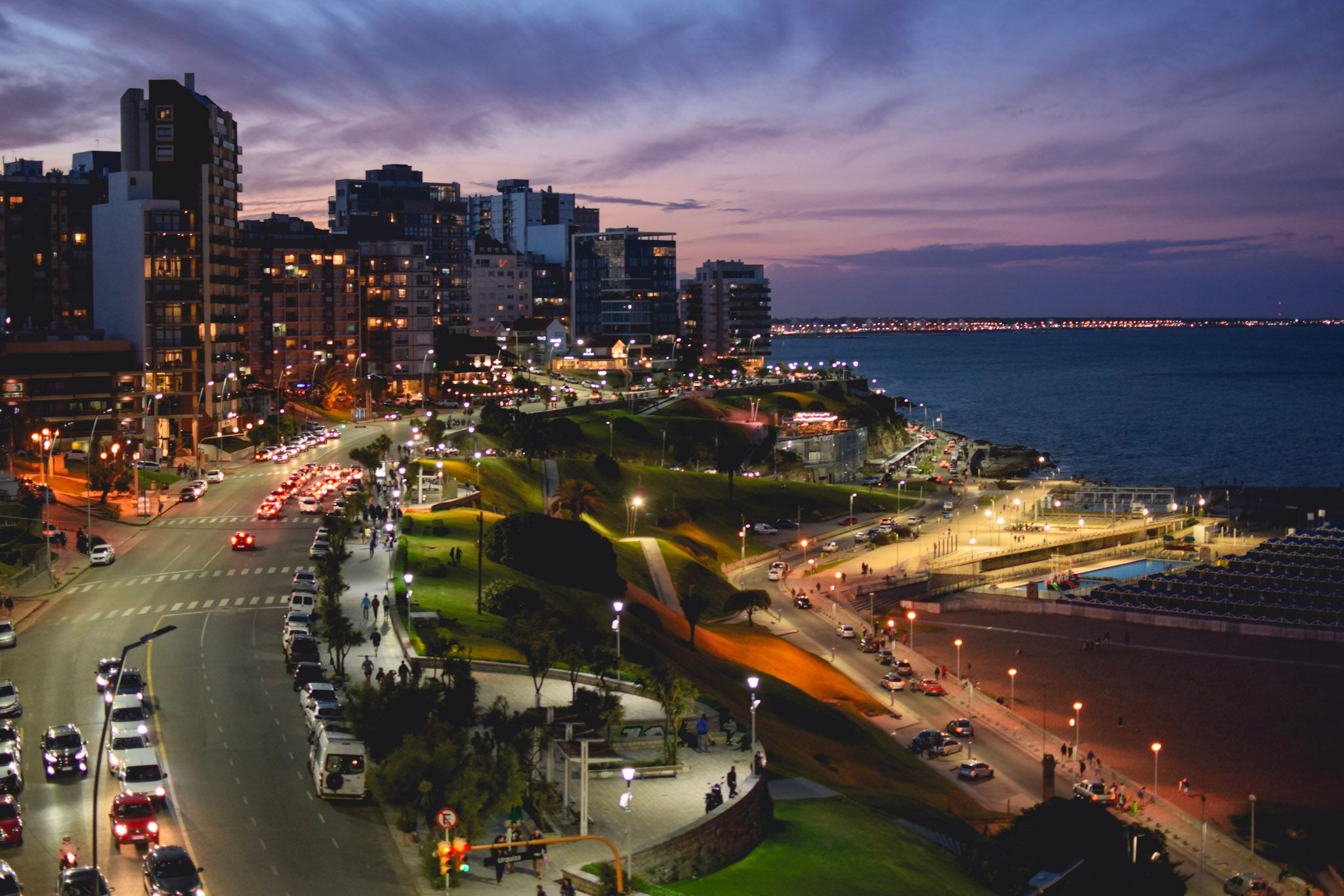 La costa de Mar del Plata de noche vista desde lo alto, con edificios iluminados, la avenida con autos y el mar bajo un cielo violeta