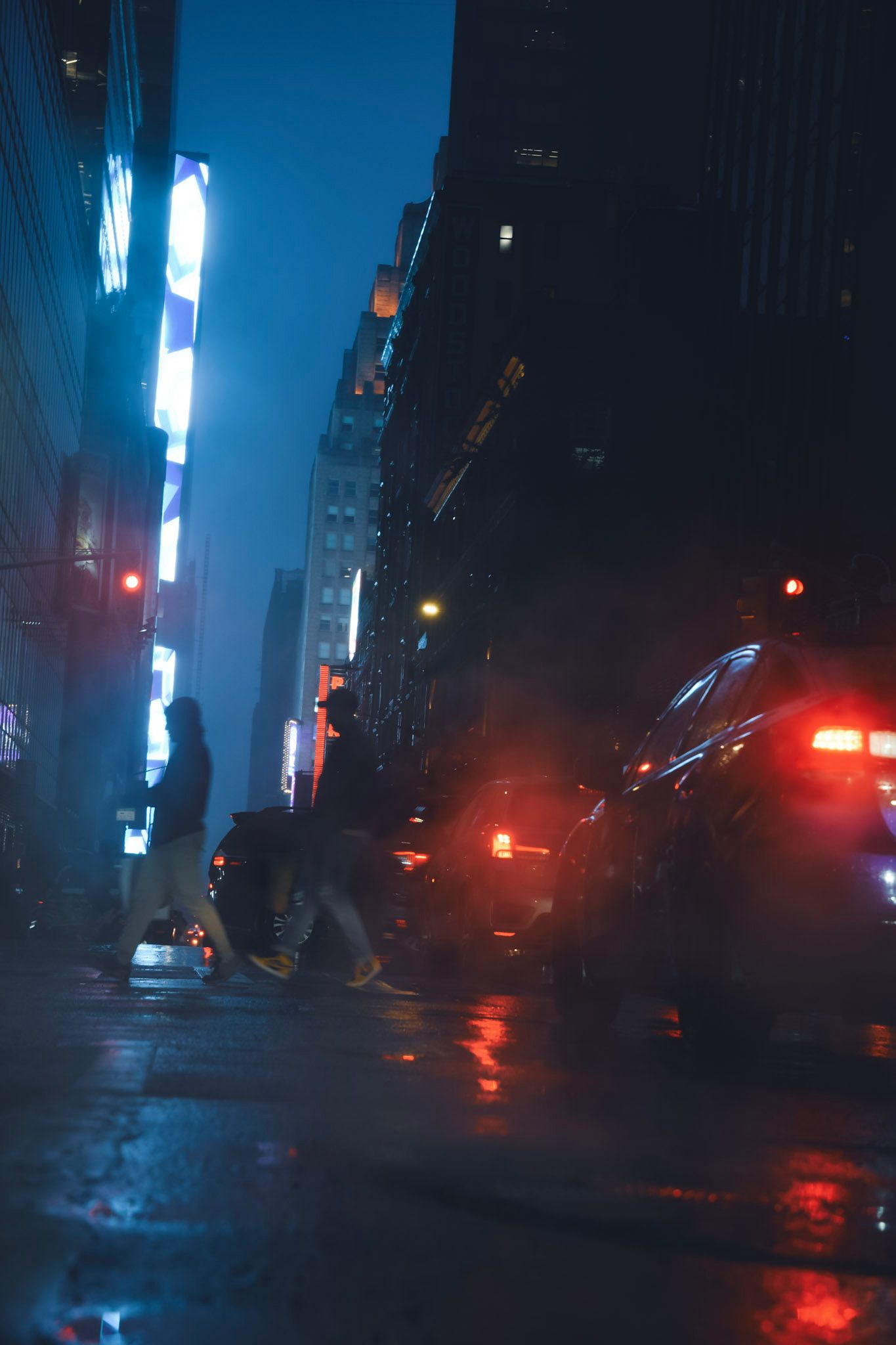 Gente cruzando una calle mojada en la penumbra azul, con las luces rojas de un auto y vapor