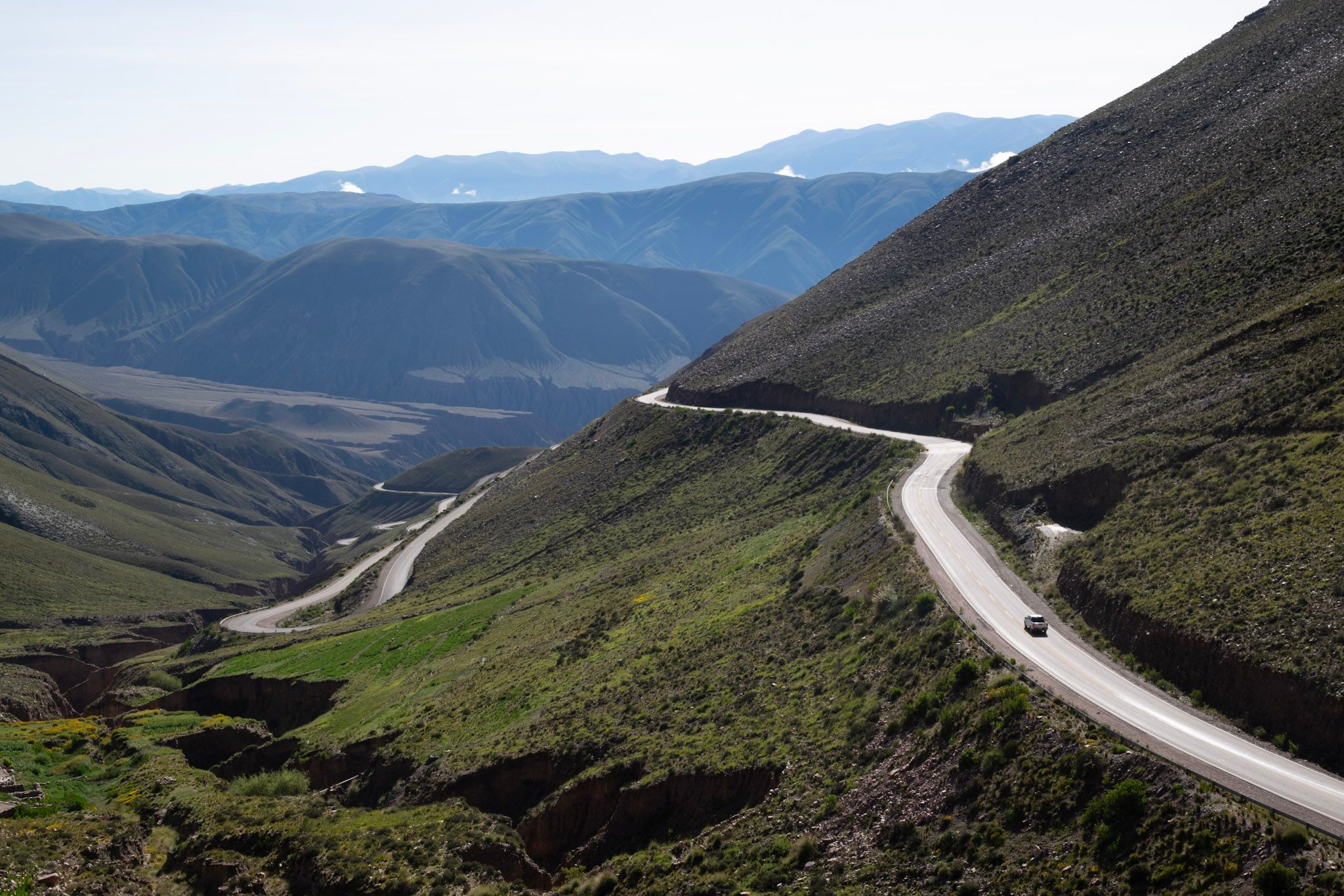 Ruta de cornisa que serpentea por la ladera verde de una quebrada, con un auto subiendo y montañas de fondo