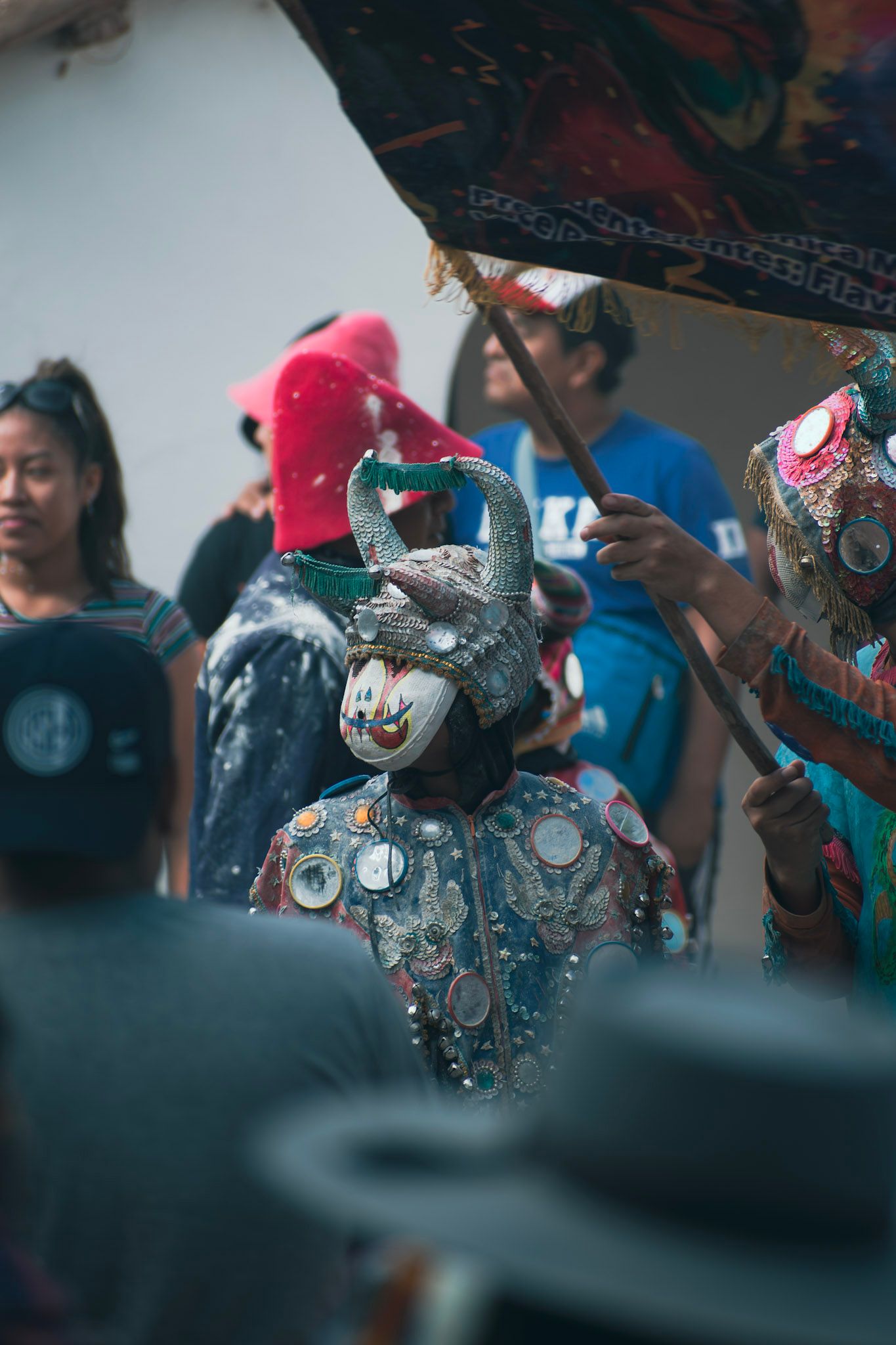 Diablo de carnaval con máscara de cuernos y traje bordado avanzando entre la gente bajo una bandera