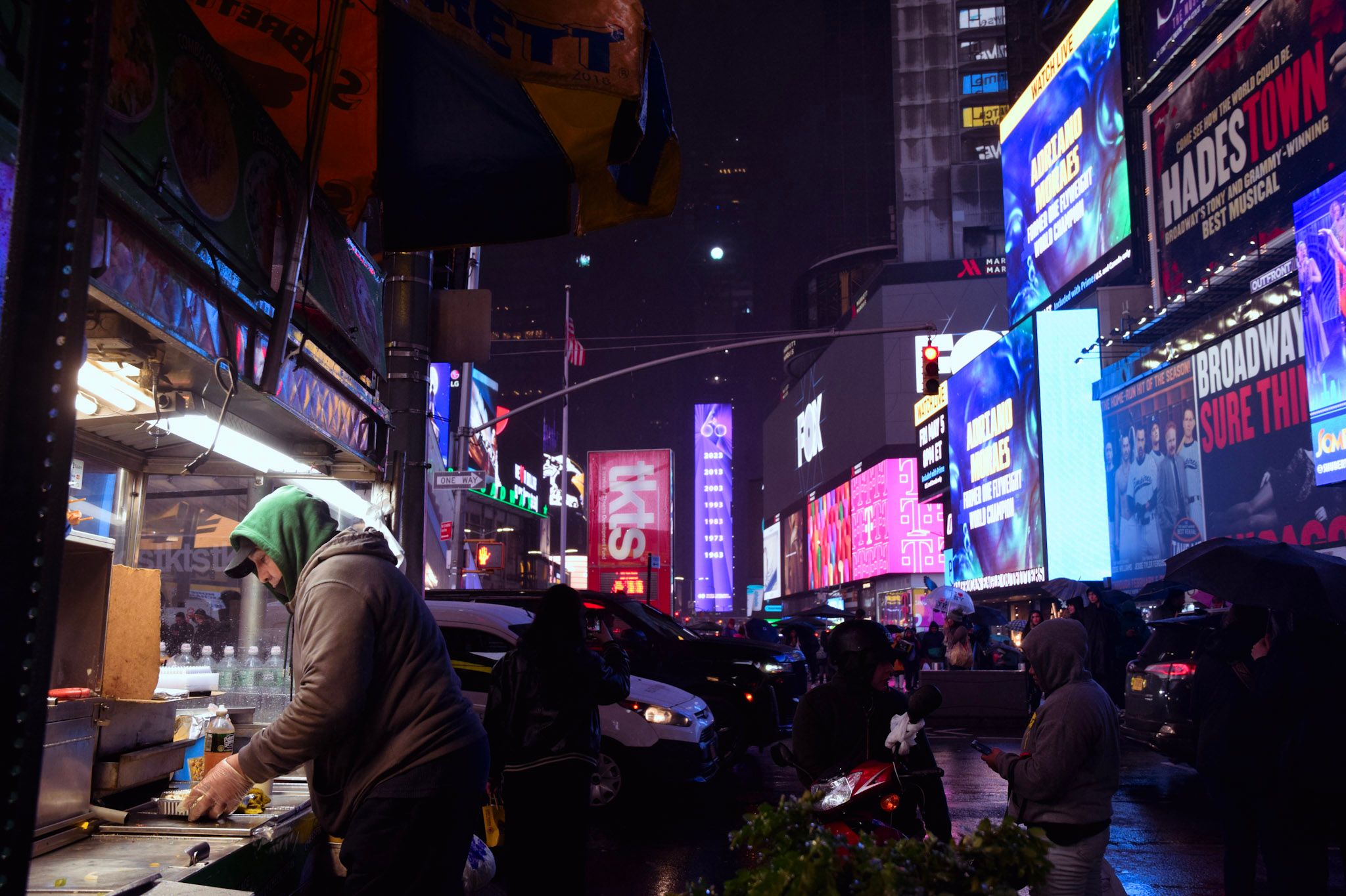 Vendedor con capucha cocinando en un carrito de comida de noche, bajo la lluvia, con las pantallas de Times Square detrás