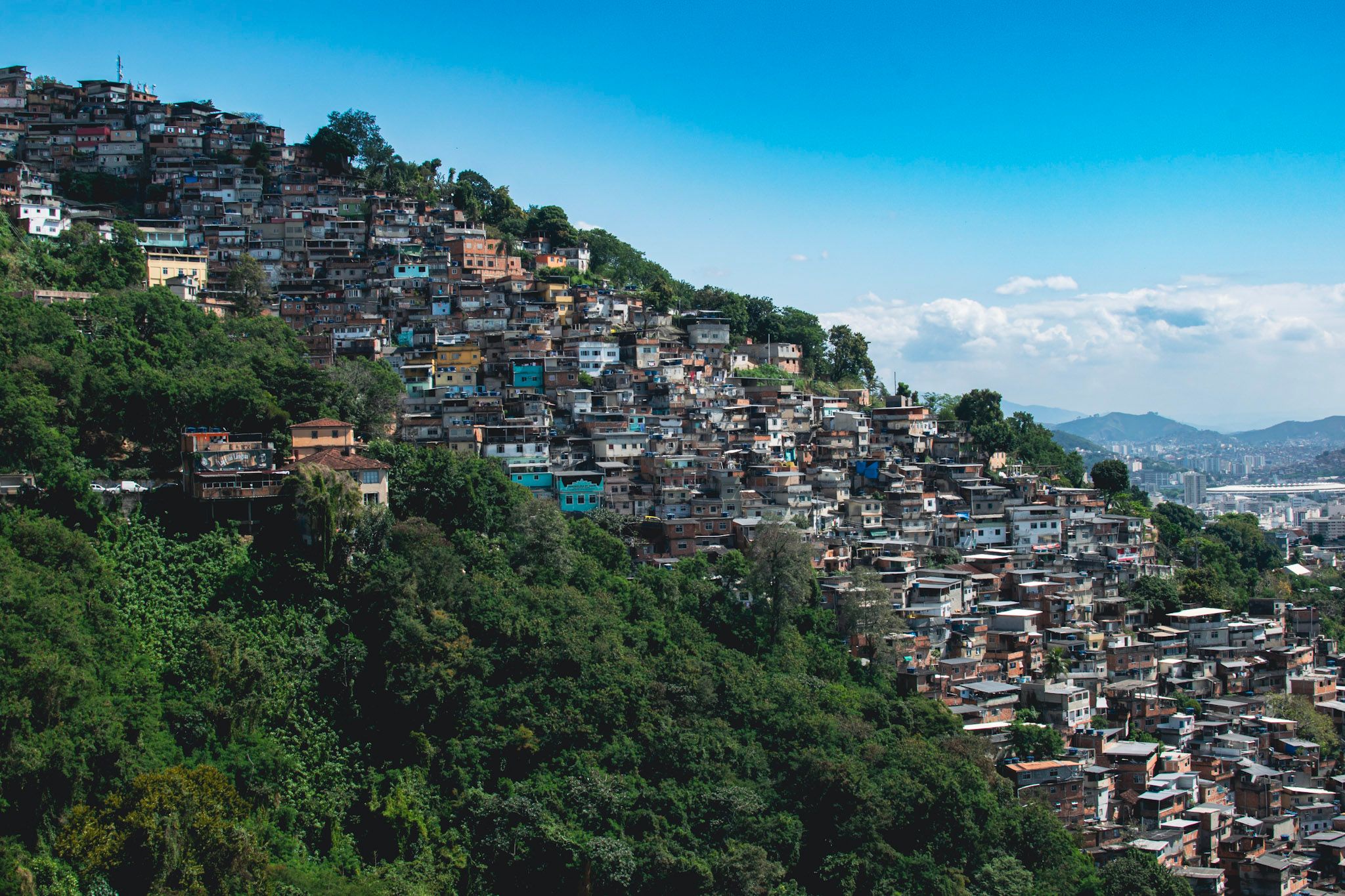Un morro cubierto de casas sobre una ladera de selva, con la ciudad y montañas al fondo bajo el cielo azul