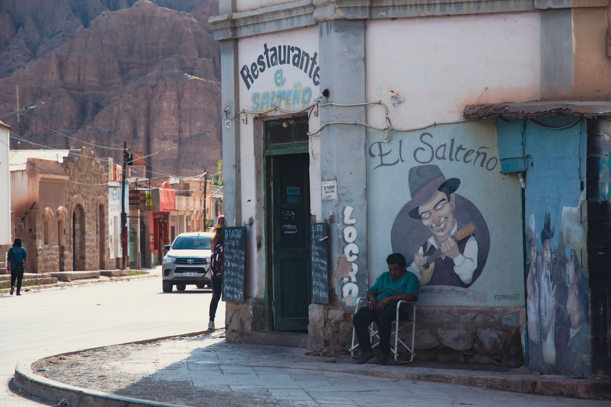 Esquina con el restaurante El Salteño, un mural de un hombre con sombrero tocando un instrumento y un señor sentado en la vereda, con cerros rojizos detrás