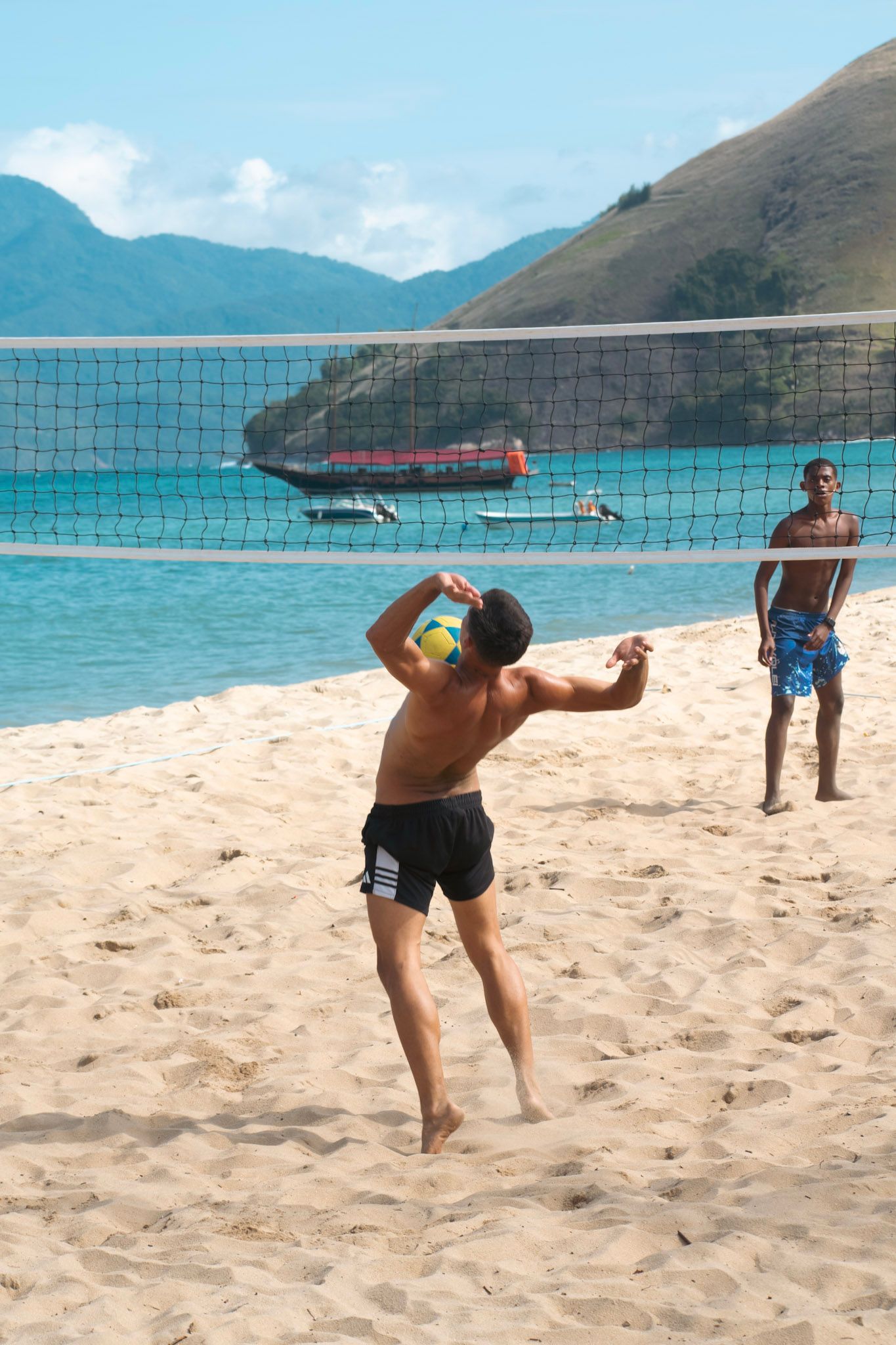 Chico de espaldas sacando con la pelota de futevóley junto a la red, en una playa con barcos y cerros
