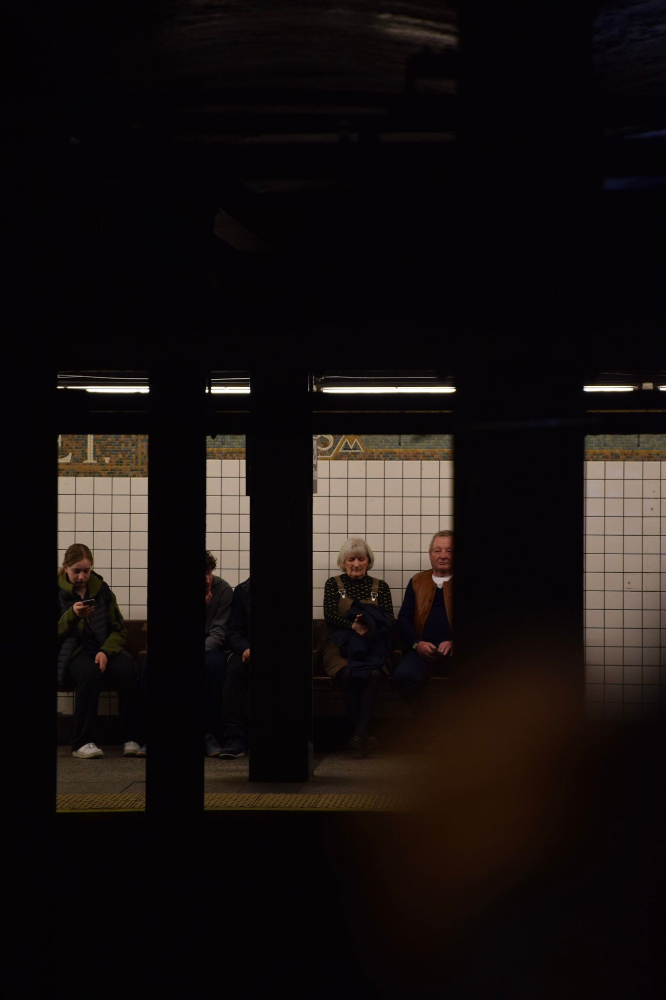 Personas sentadas en un banco del andén del subte, vistas entre columnas oscuras desde el otro lado de las vías