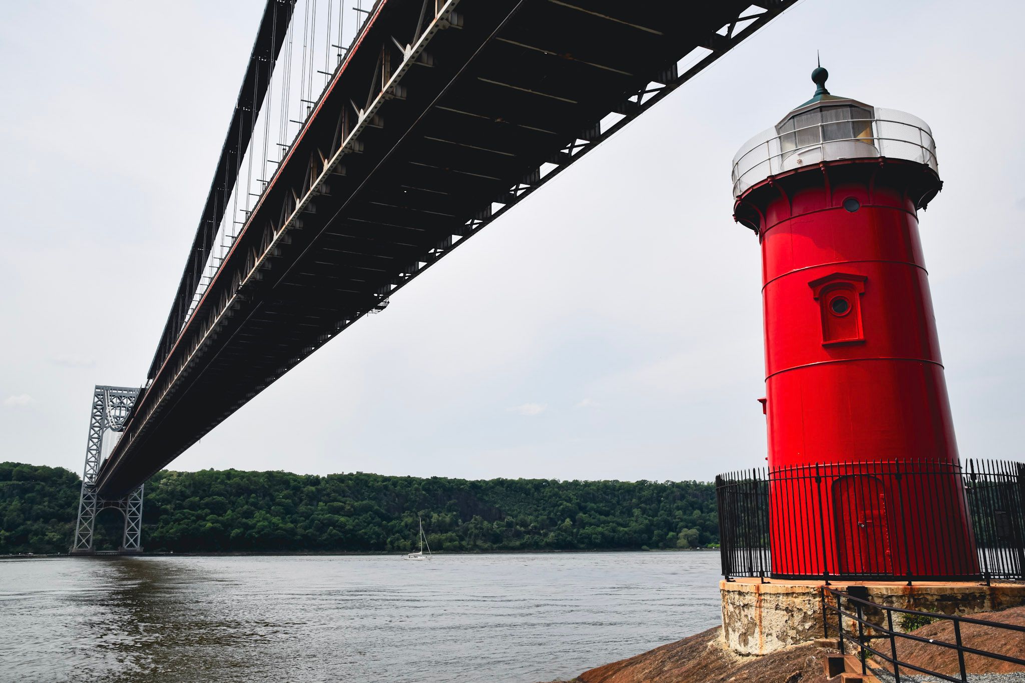 El faro rojo de Jeffrey's Hook al pie del puente George Washington, visto desde abajo, con el río Hudson y la costa arbolada de fondo