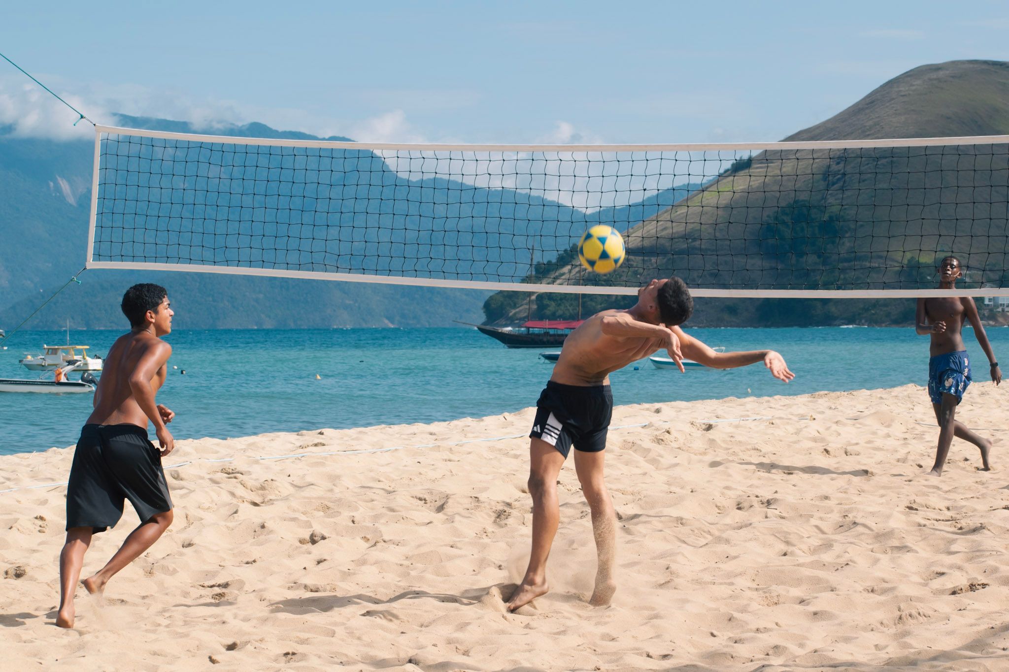 Chicos jugando al futevóley en la playa, uno a punto de cabecear la pelota, con el mar y cerros de fondo