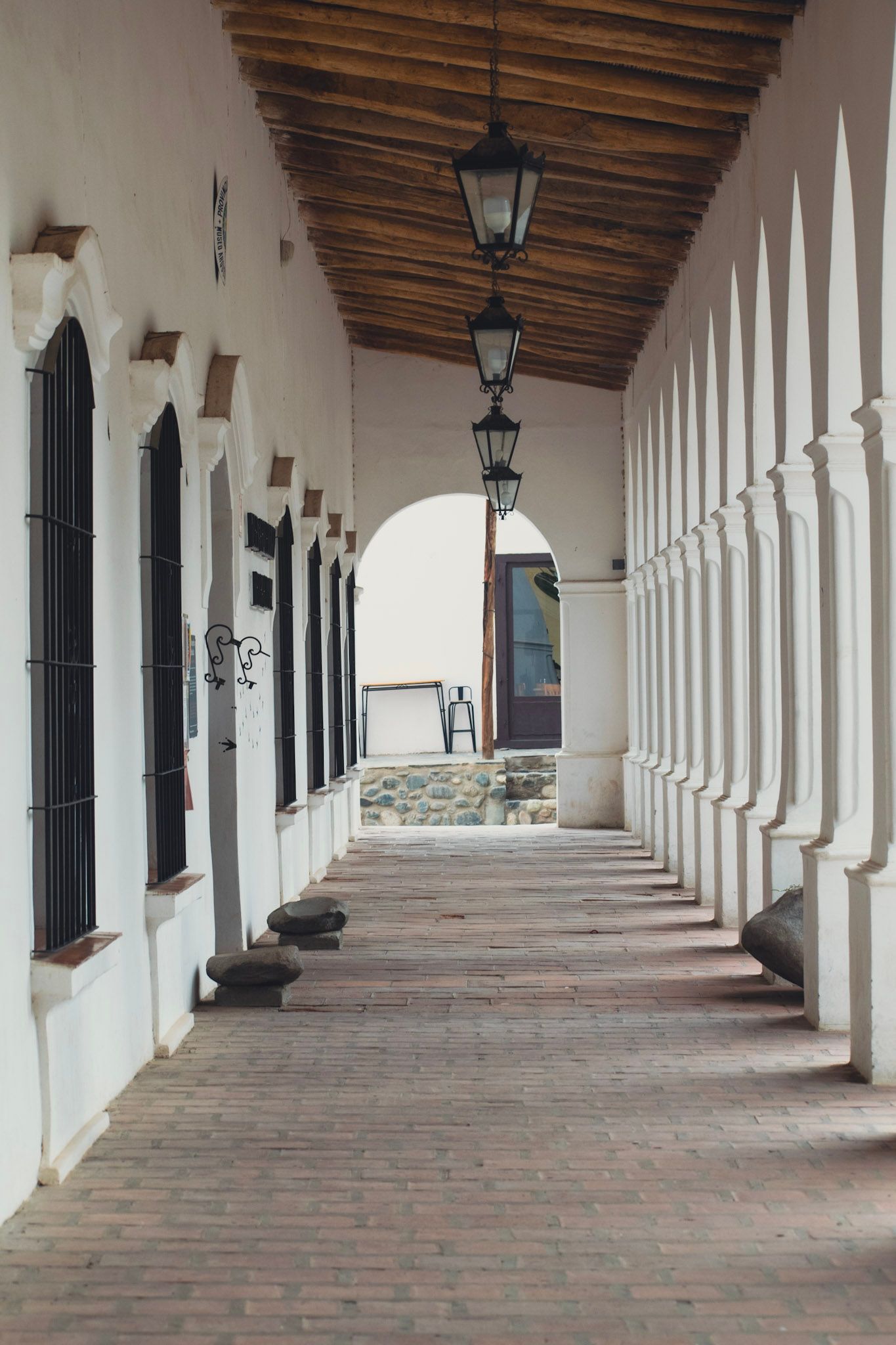 Galería colonial blanca con arcos, faroles colgando de un techo de madera y piso de ladrillo