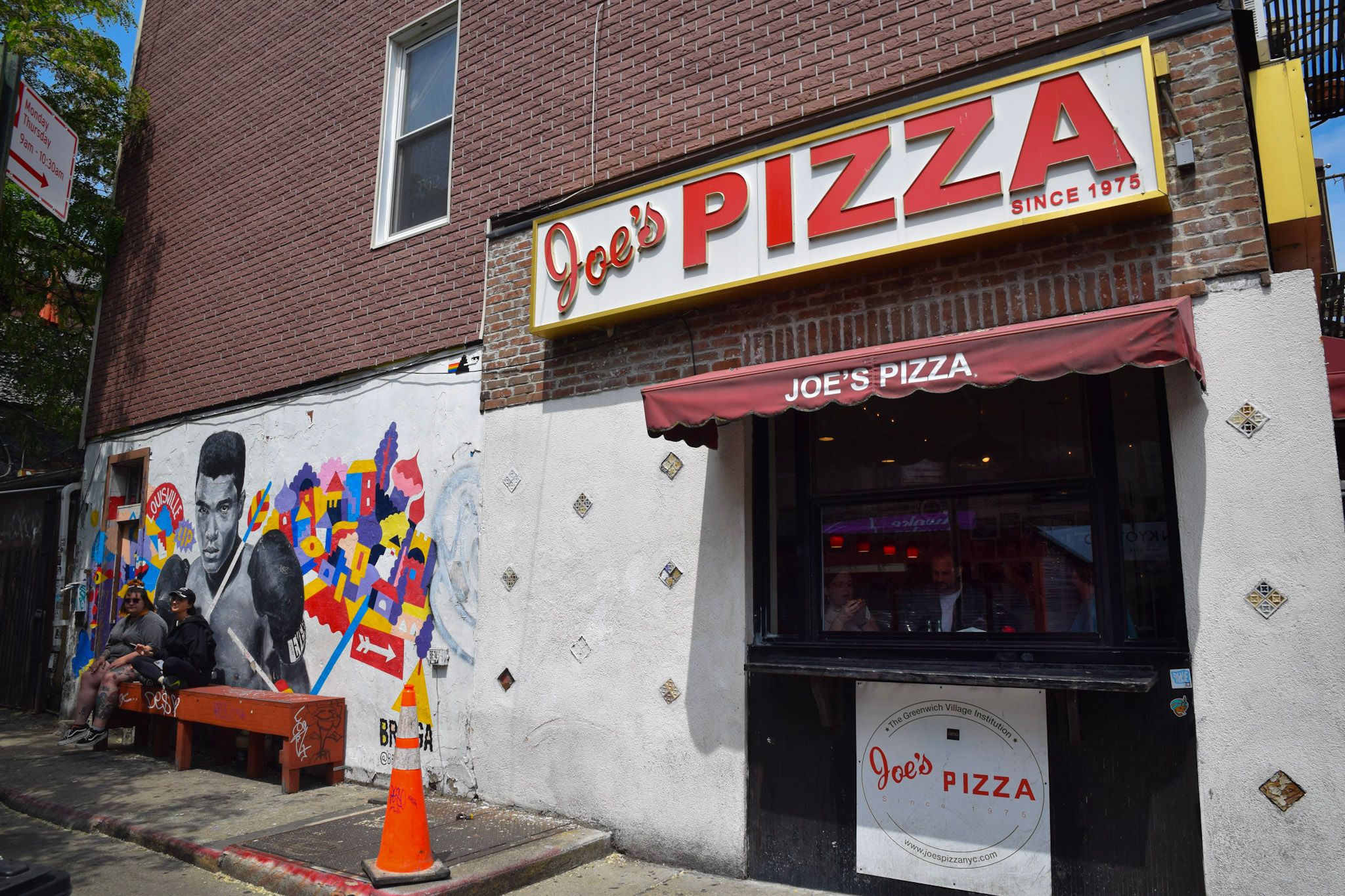 Fachada de Joe's Pizza con un mural de un boxeador y figuras de colores en la pared lateral, y gente sentada en un banco