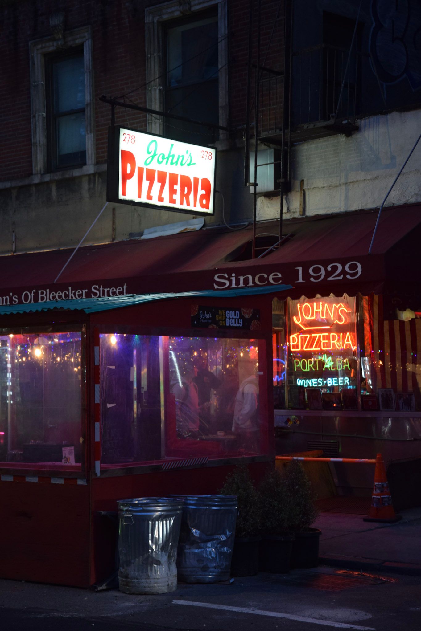 Fachada de John's Pizzeria de noche, con carteles de neón rojo y dos tachos de basura de metal en la vereda