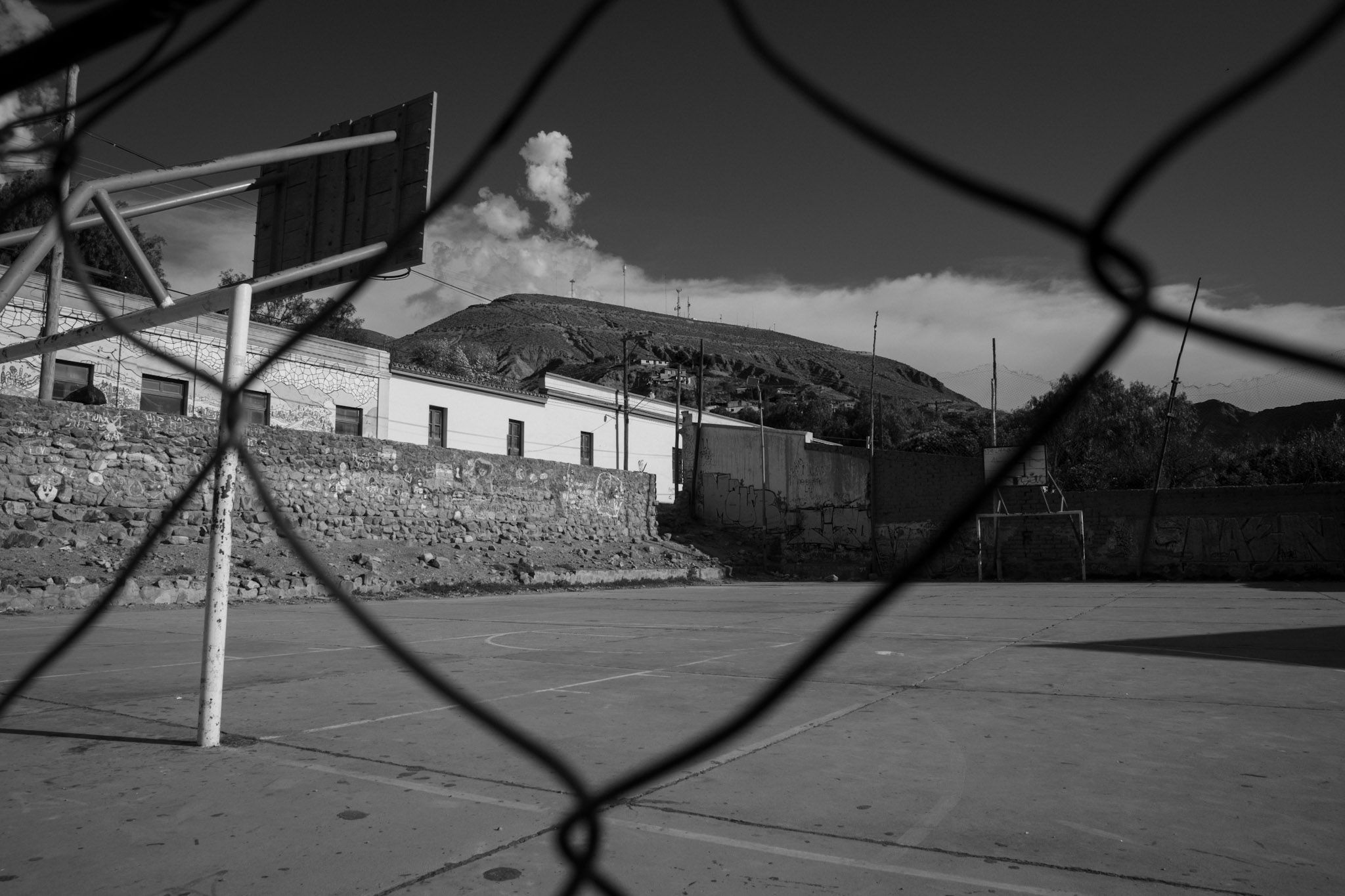 Cancha de cemento con un aro de básquet y cerros de fondo, vista a través de un alambrado, en blanco y negro