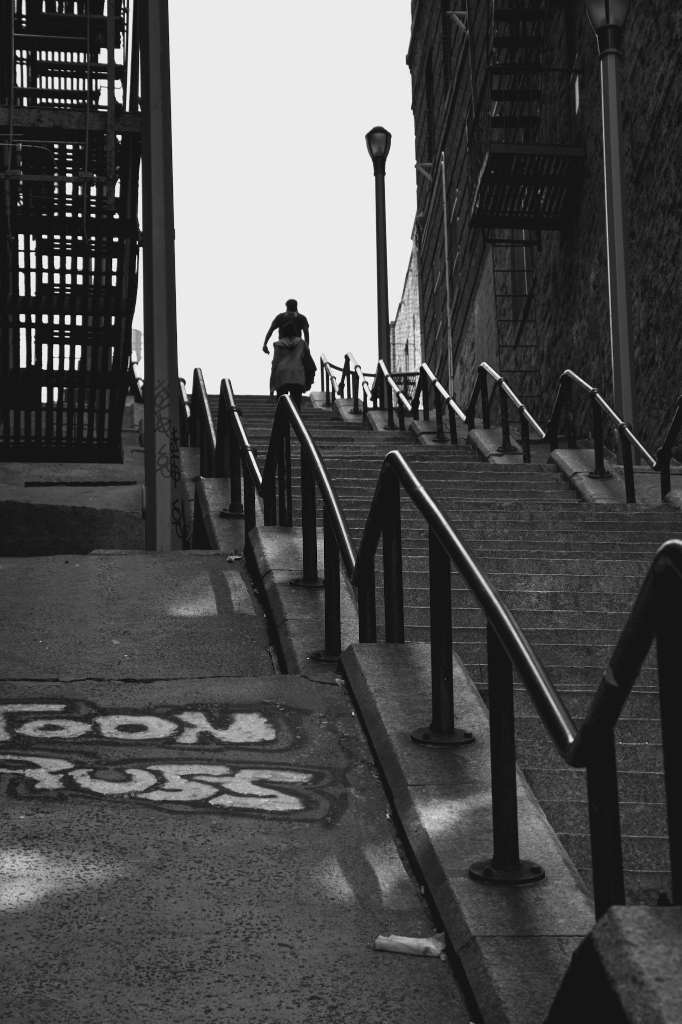 Una persona en silueta subiendo una larga escalera urbana con barandas, entre edificios, en blanco y negro
