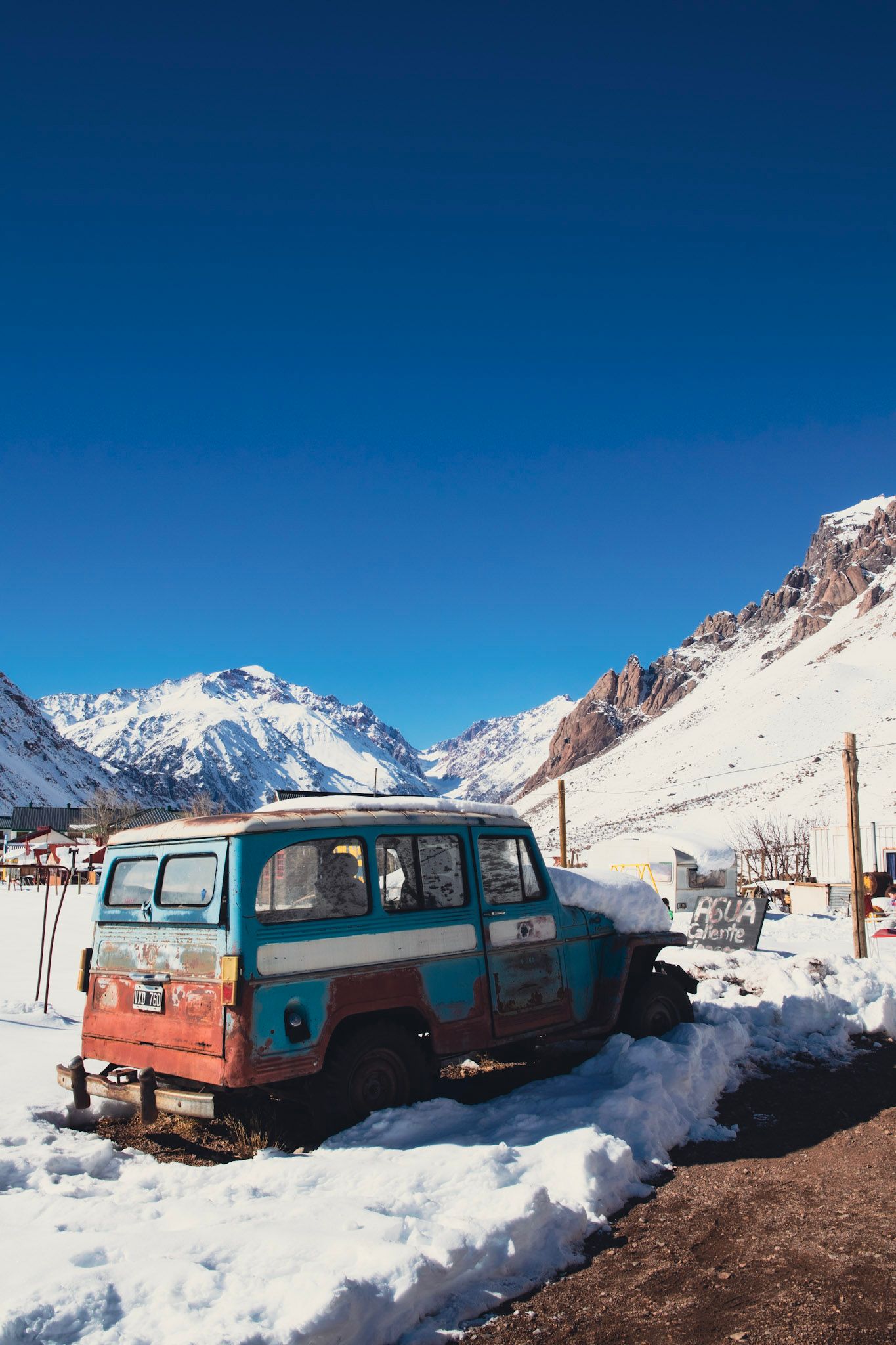 Camioneta antigua celeste y oxidada estacionada en la nieve, con montañas nevadas y cielo azul profundo