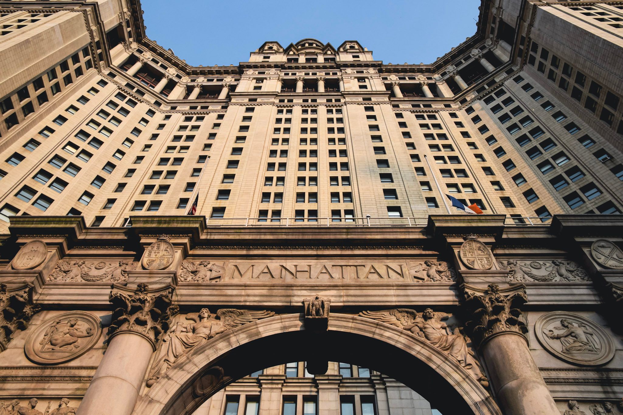 El Manhattan Municipal Building visto desde abajo, con el arco de piedra tallada y la palabra MANHATTAN, iluminado por el sol de la tarde