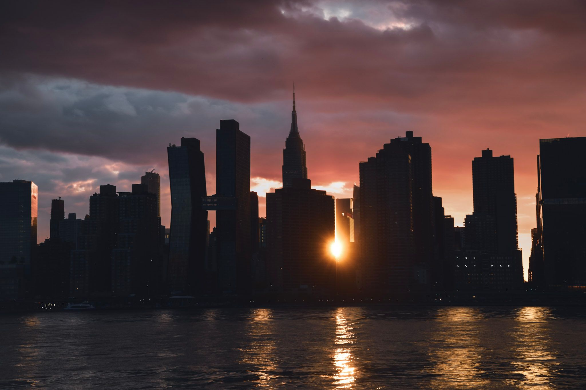El skyline de Manhattan en silueta al atardecer, con el sol asomando entre los edificios y reflejándose en el río bajo nubes rojizas
