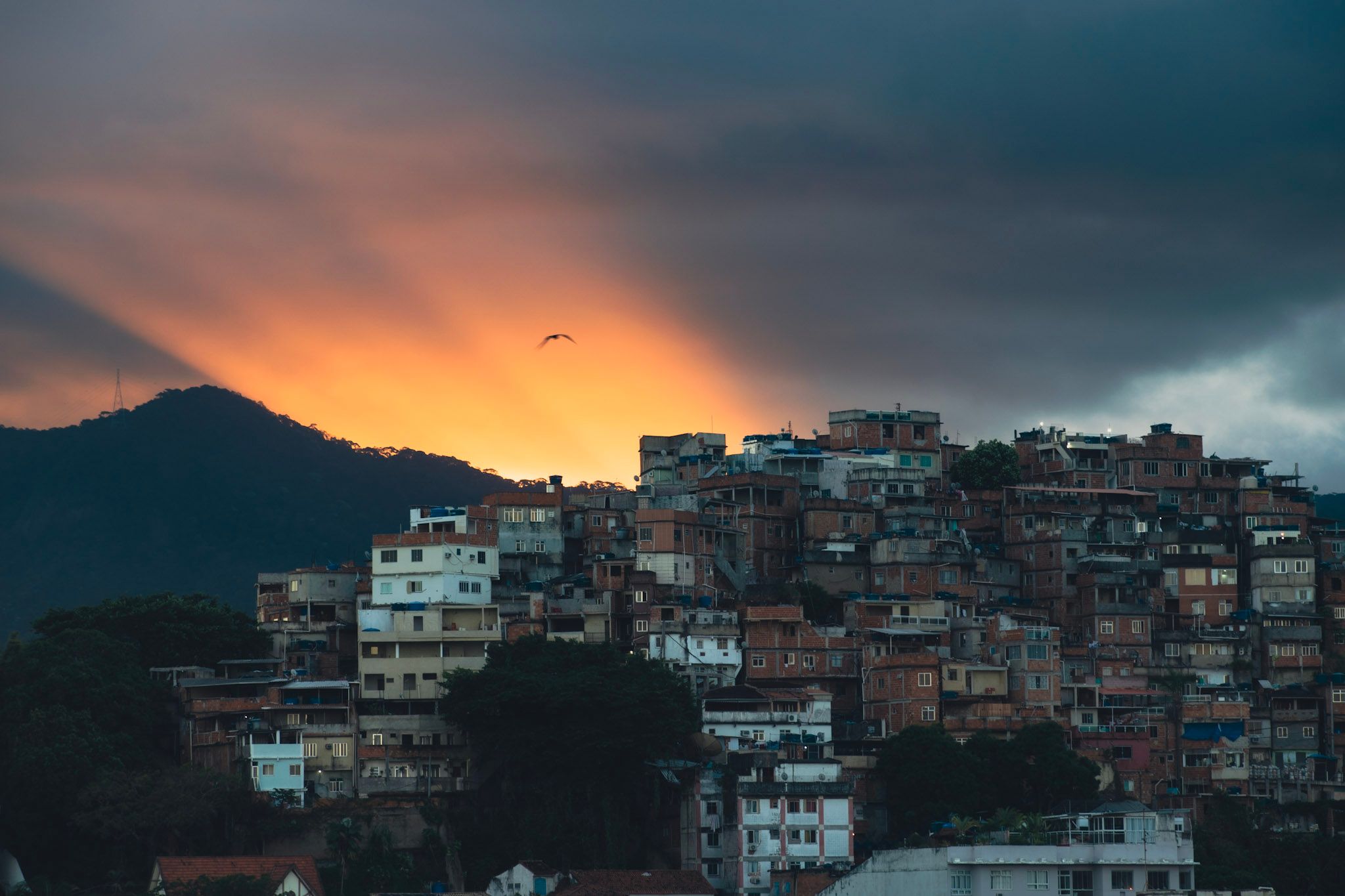 Casas de un morro apiñadas en la ladera bajo un cielo oscuro atravesado por rayos de sol anaranjados