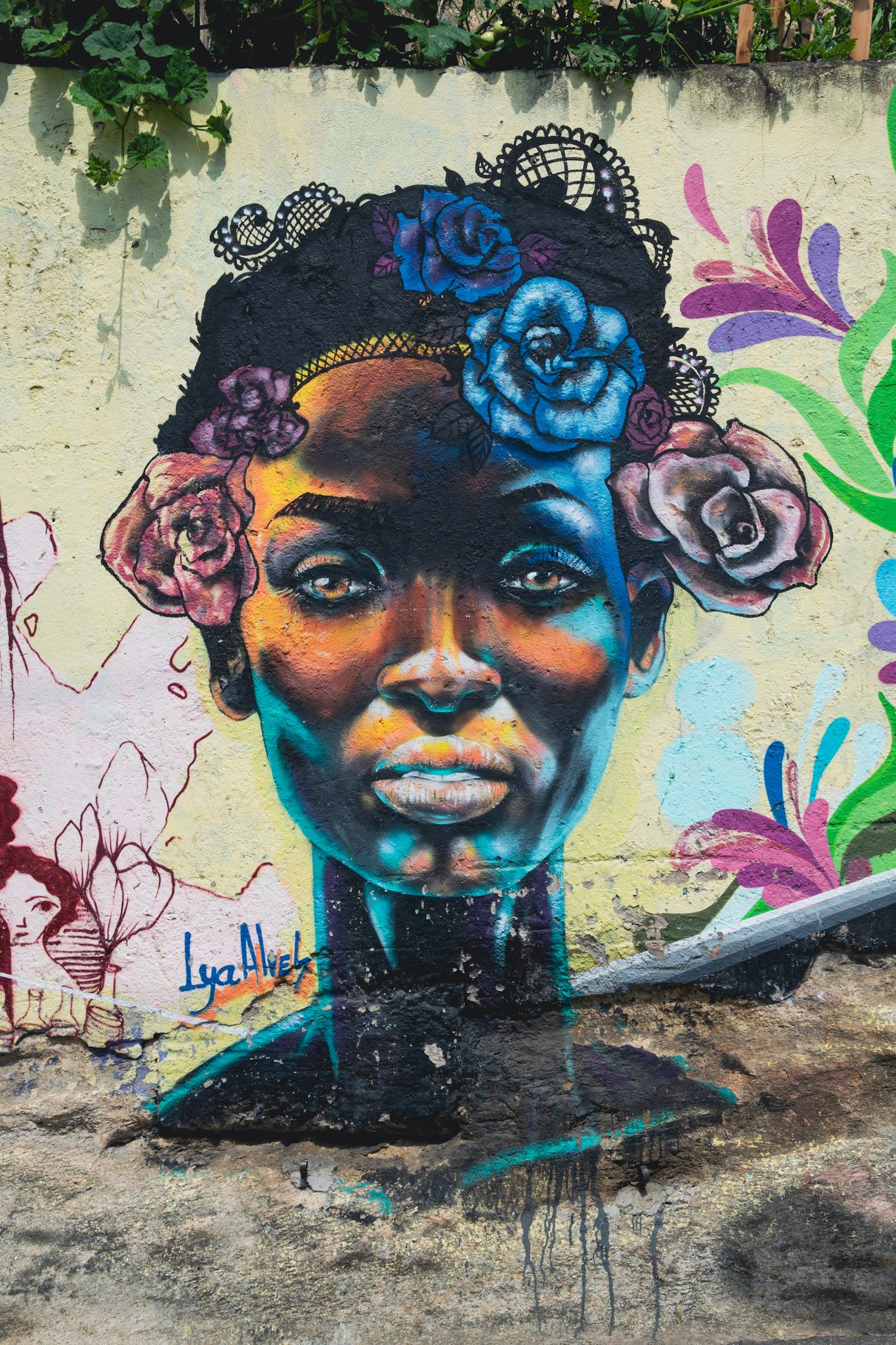 Mural del rostro de una mujer con flores azules y rosadas en el pelo, pintado en un muro