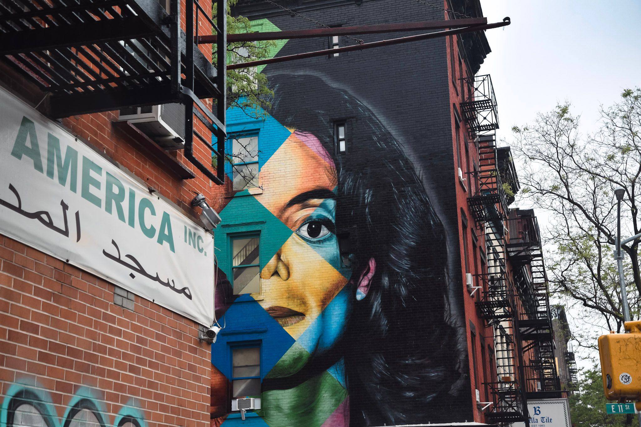 Mural de un rostro de mujer con pelo negro y colores geométricos, entre escaleras de incendio y un cartel en inglés y árabe