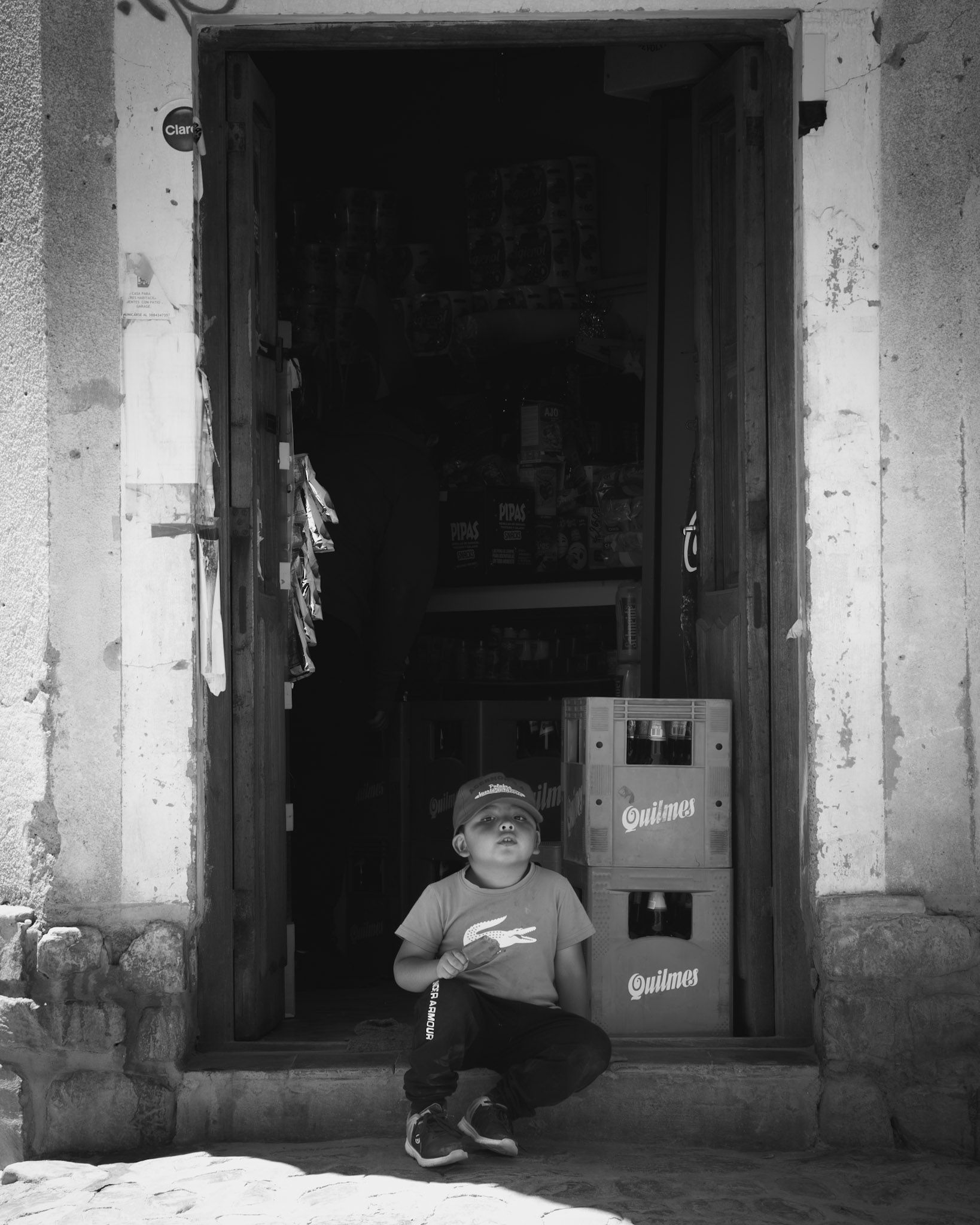 Nene en cuclillas con gorra en la puerta de un almacén, junto a cajones de cerveza, en blanco y negro