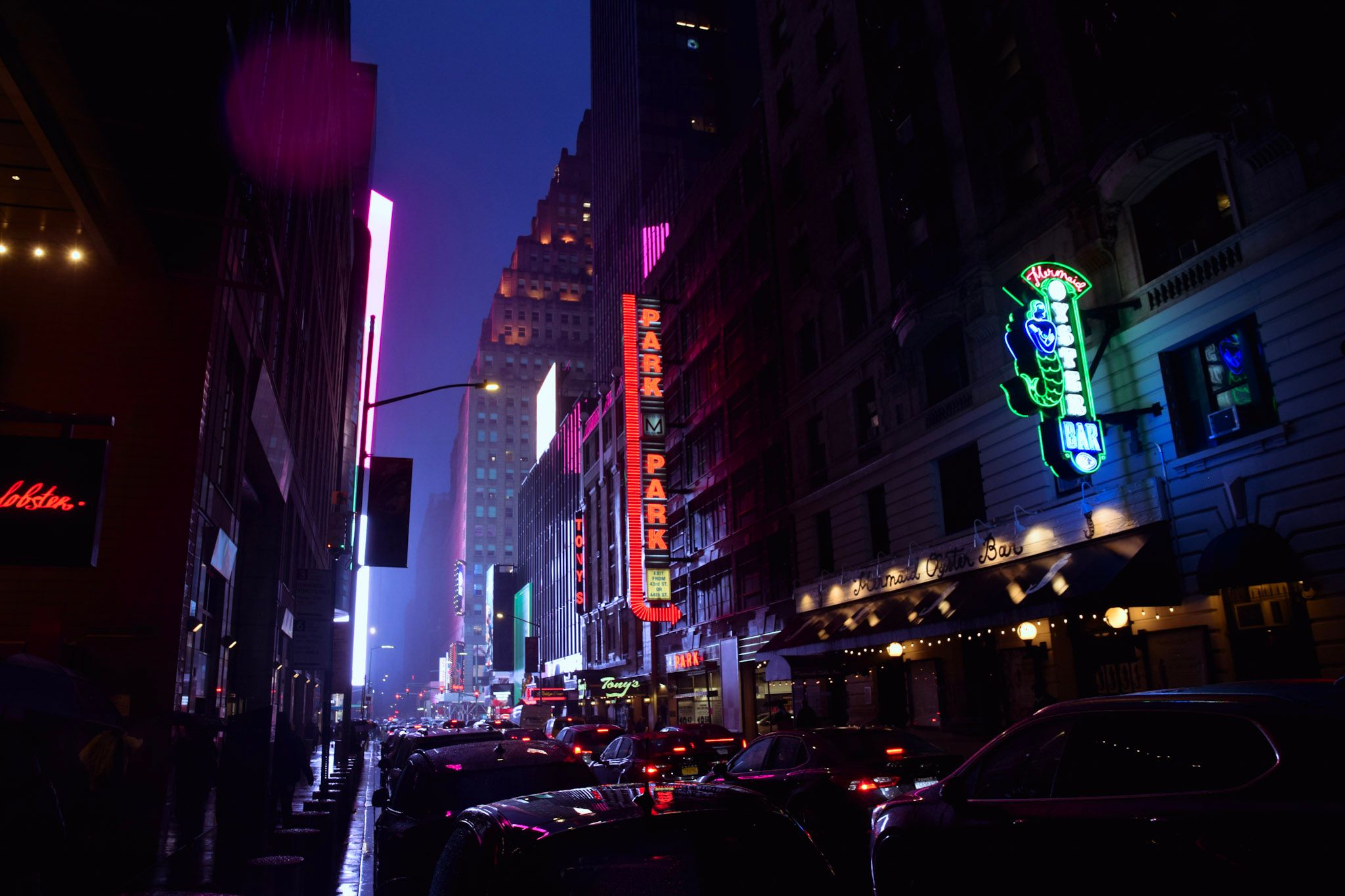 Calle oscura con autos estacionados y carteles de neón rojo y verde, bajo un cielo azul de noche
