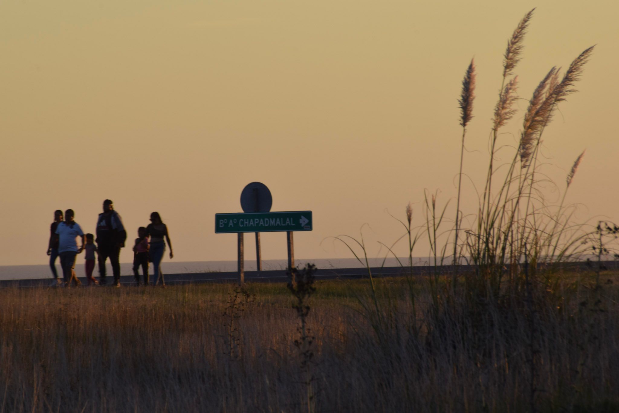 Siluetas de una familia caminando al atardecer junto a un cartel de ruta y cortaderas, bajo un cielo anaranjado