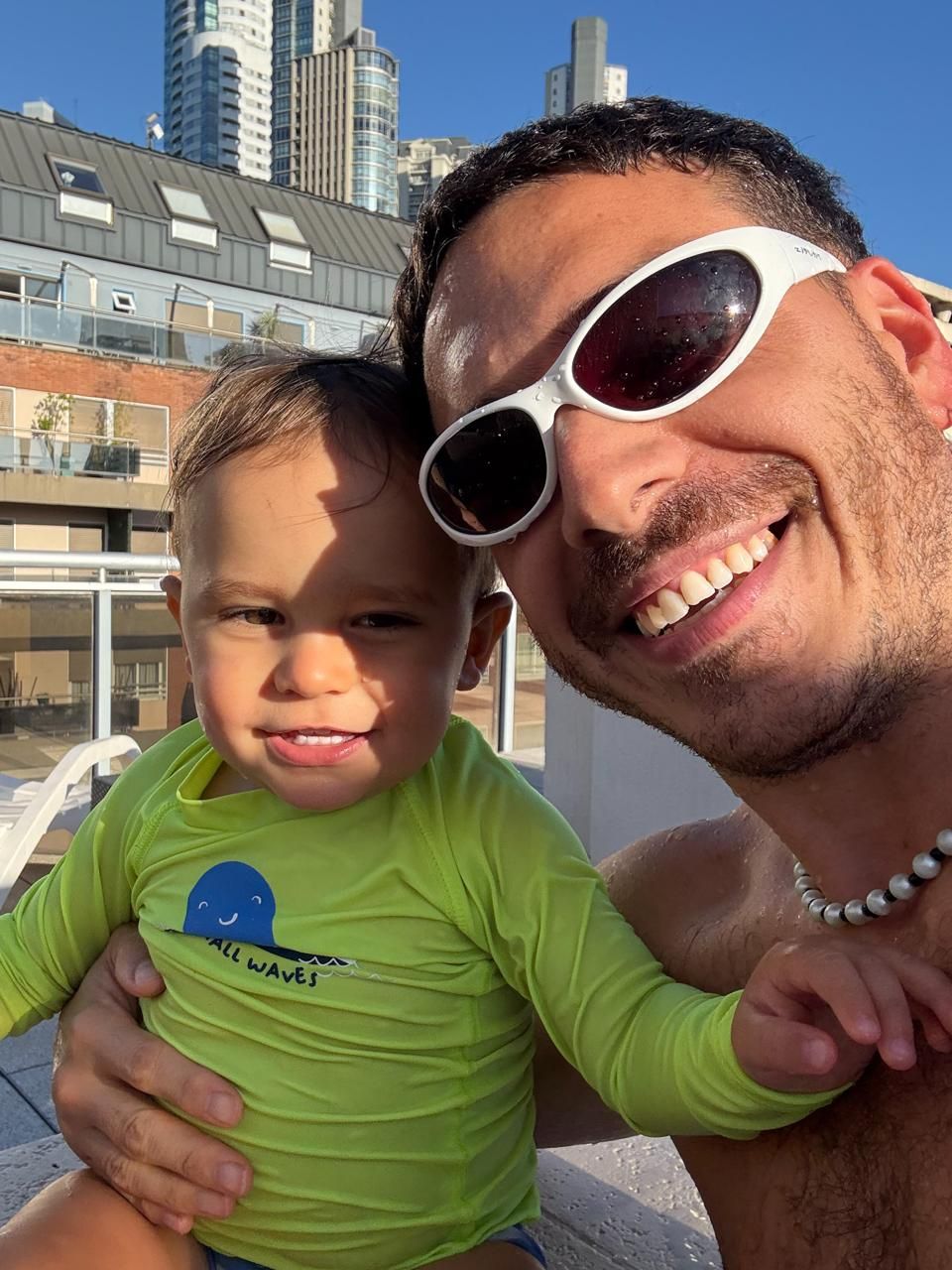 Mauruch con anteojos de sol blancos, sonriendo junto a su hijo en una terraza con edificios de fondo