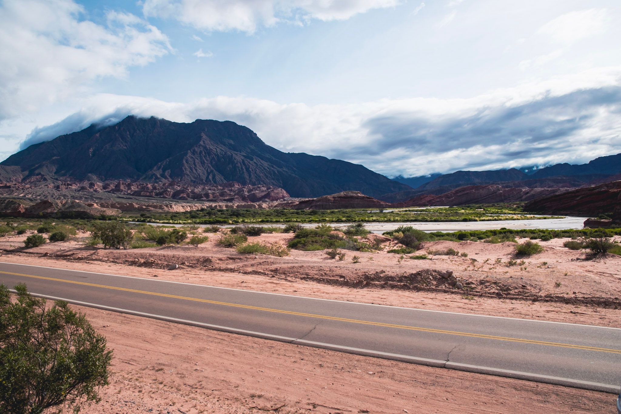 Ruta que cruza un paisaje árido con arbustos, un río y un cerro oscuro con nubes bajas