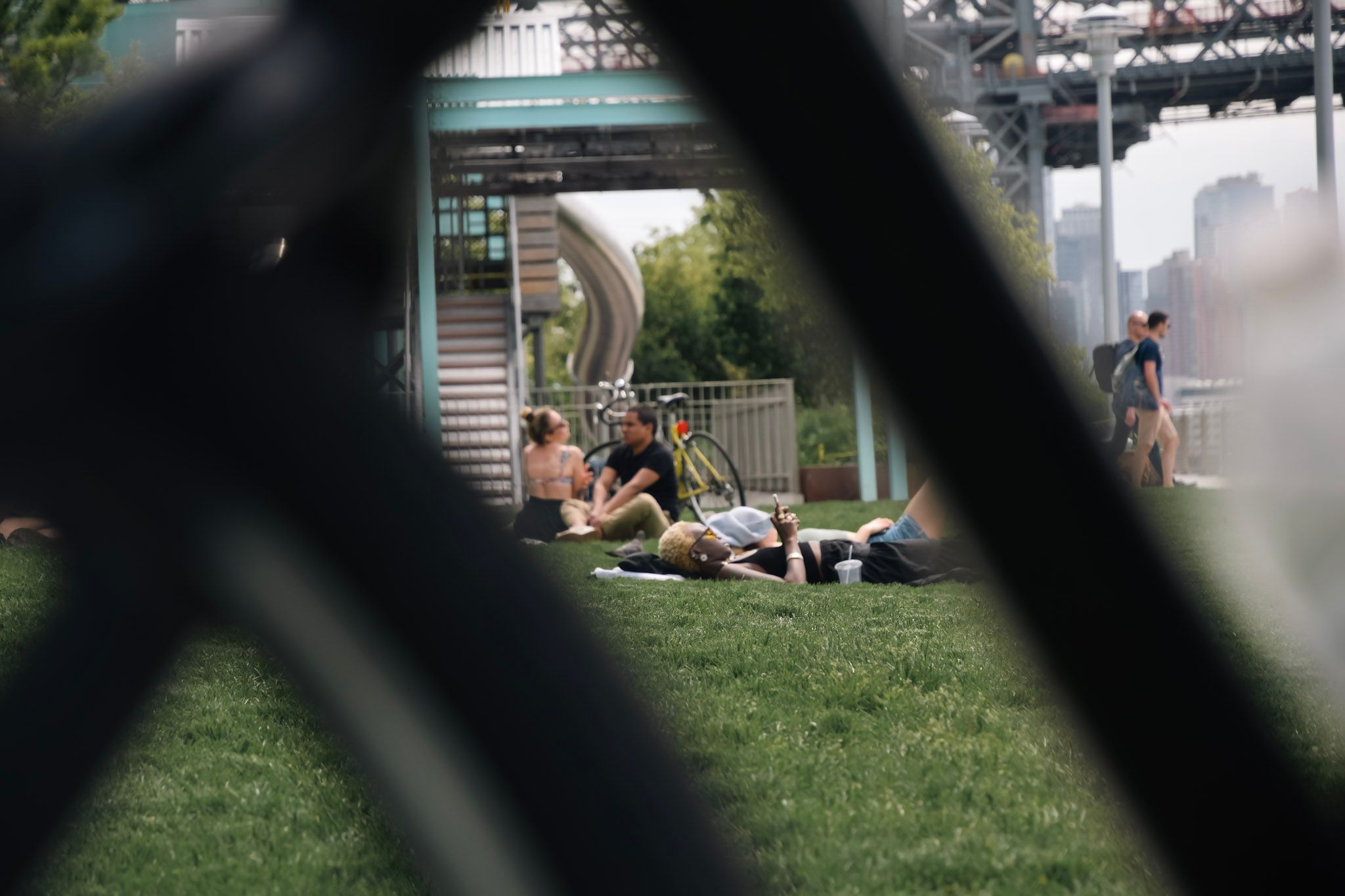Gente sentada y acostada en el pasto de un parque junto a una bicicleta, vista a través de una reja desenfocada en primer plano