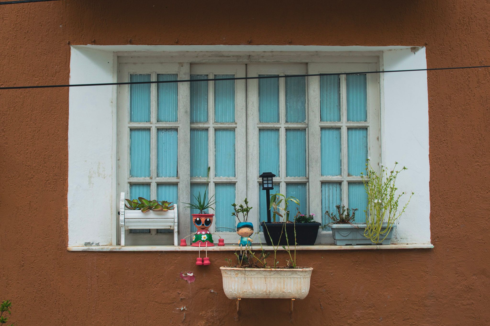 Ventana blanca con persianas celestes sobre una pared marrón, con macetas y muñequitos en el alféizar